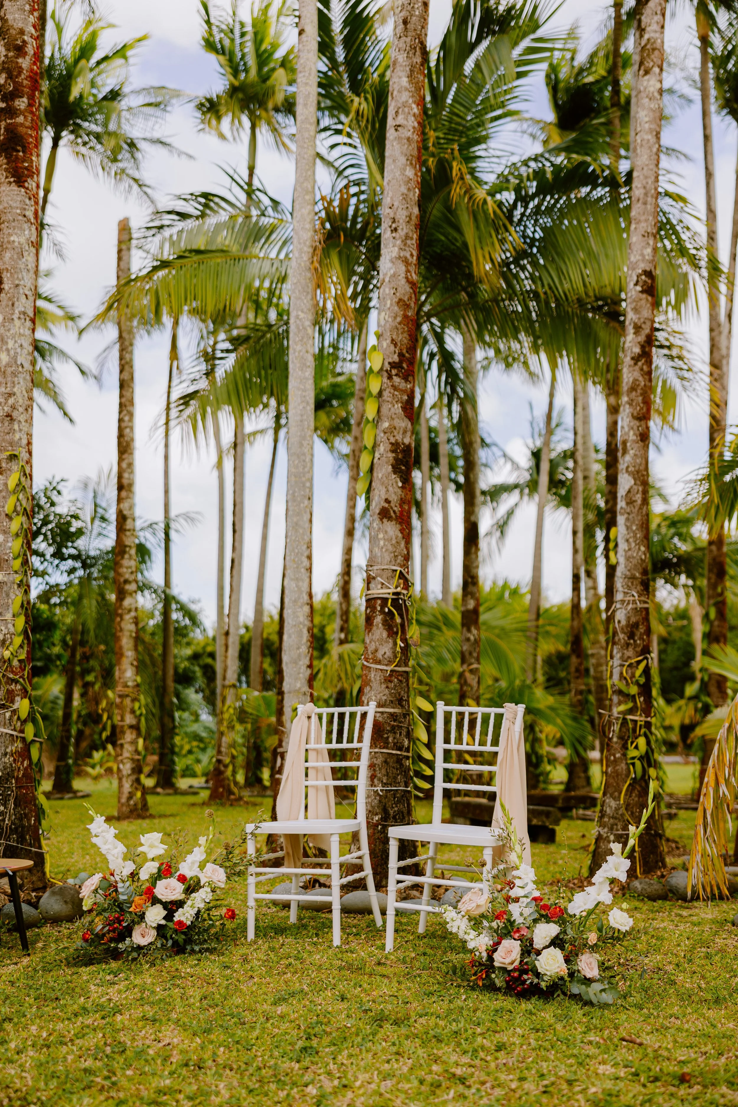 Chaises blanches sur l'herbe avec décor de fleurs en bas, dans un cadre de forêt tropicale avec des palmiers et des arbres.