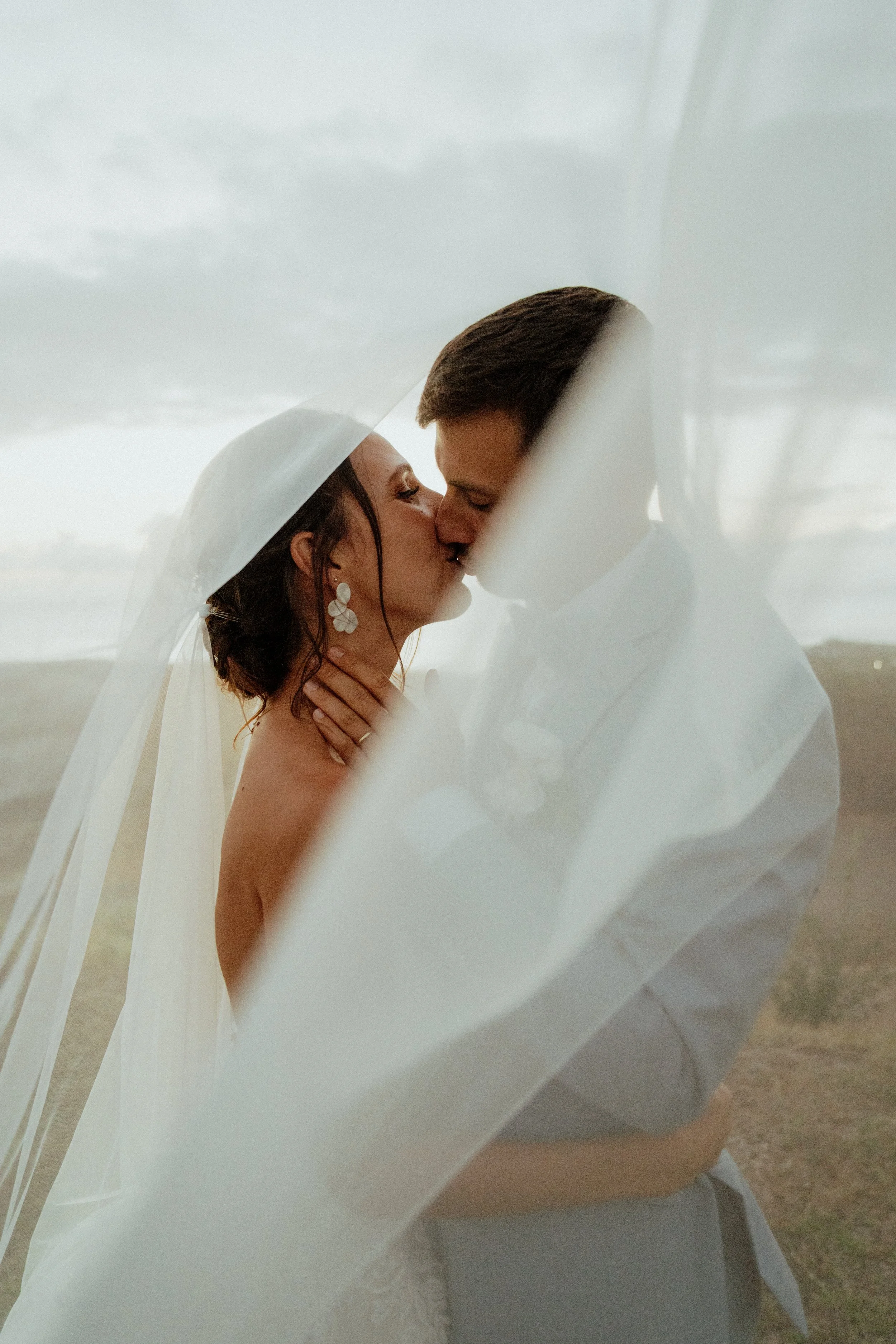 Un couple s'embrasse tendrement, la femme porte un voile de mariée et des boucles d'oreilles, dans un environnement extérieur.