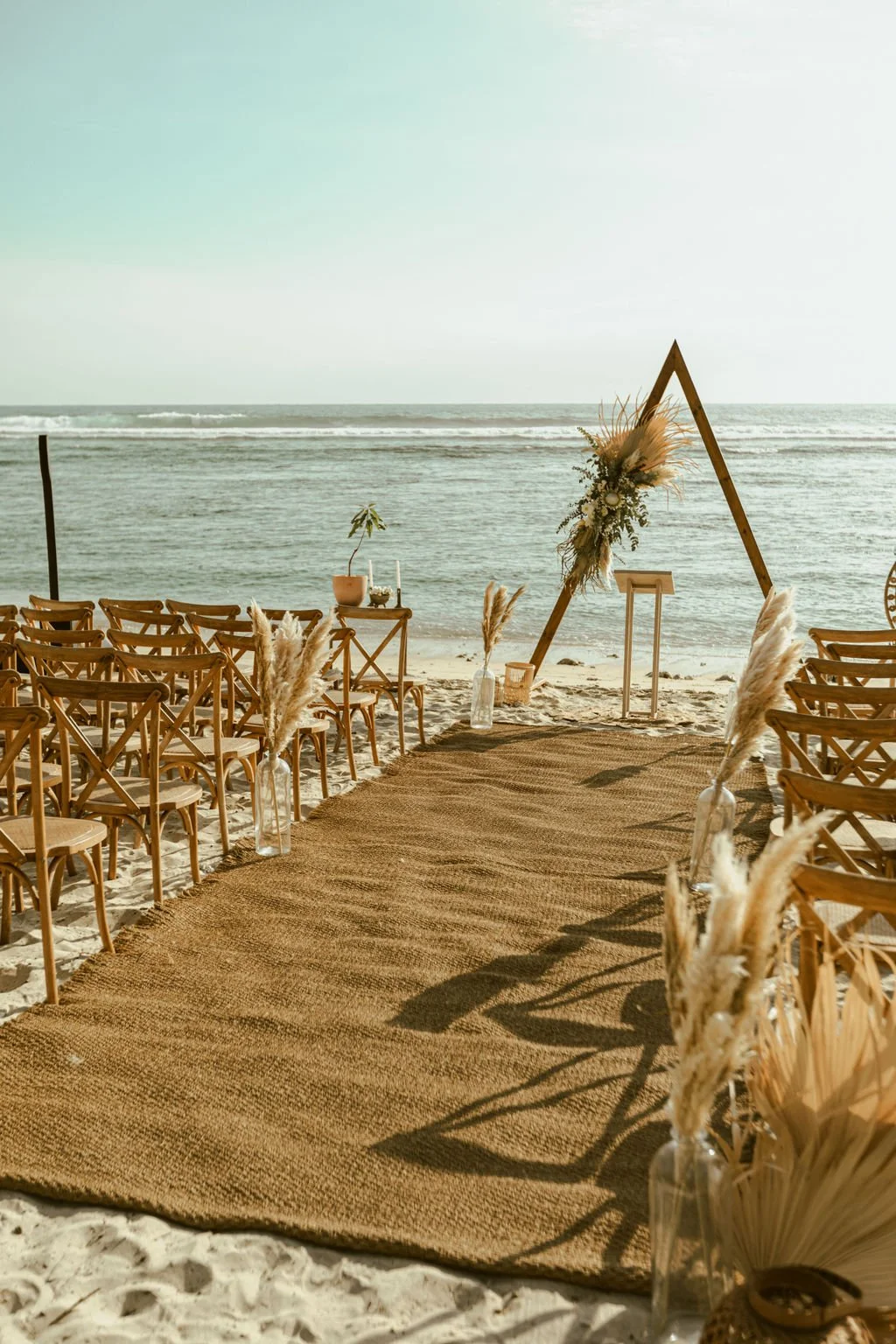 Une cérémonie de mariage en bord de mer avec un chêne en bois décoré de fleurs, des chaises en bois pour les invités, et un tapis en jute sur le sable.