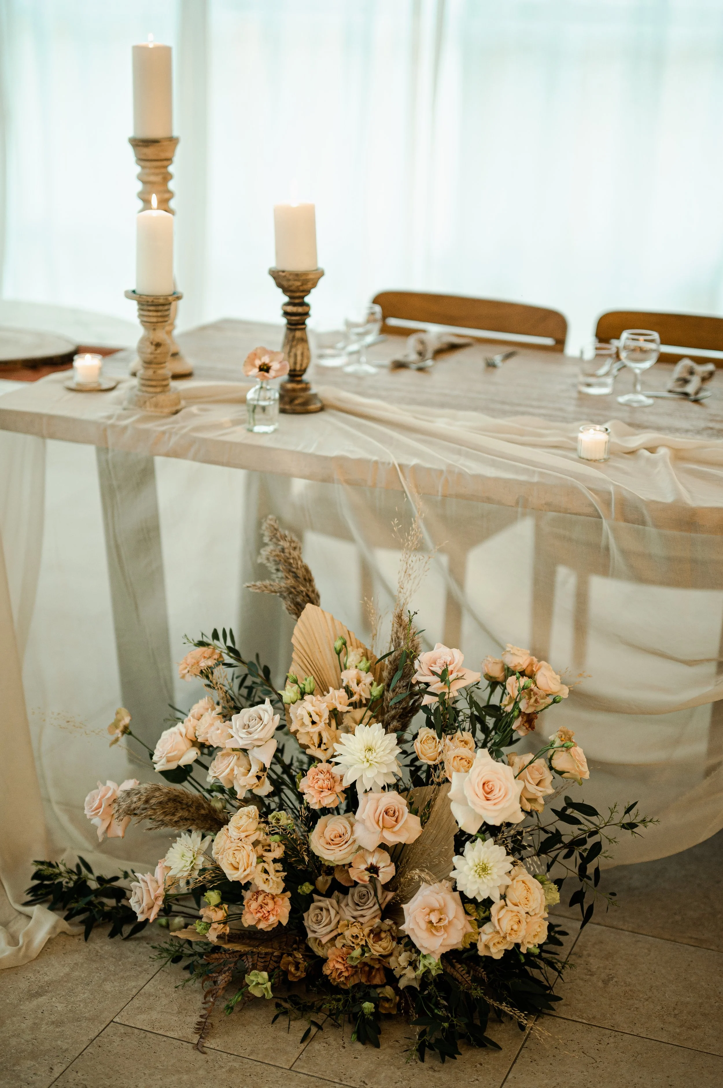 Arrangement floral avec des roses, des dahlias et des pampas, disposé dans un panier en osier. Décoré avec du tissu beige et placé sur un sol en carrelage avec un fond de rideaux blancs.