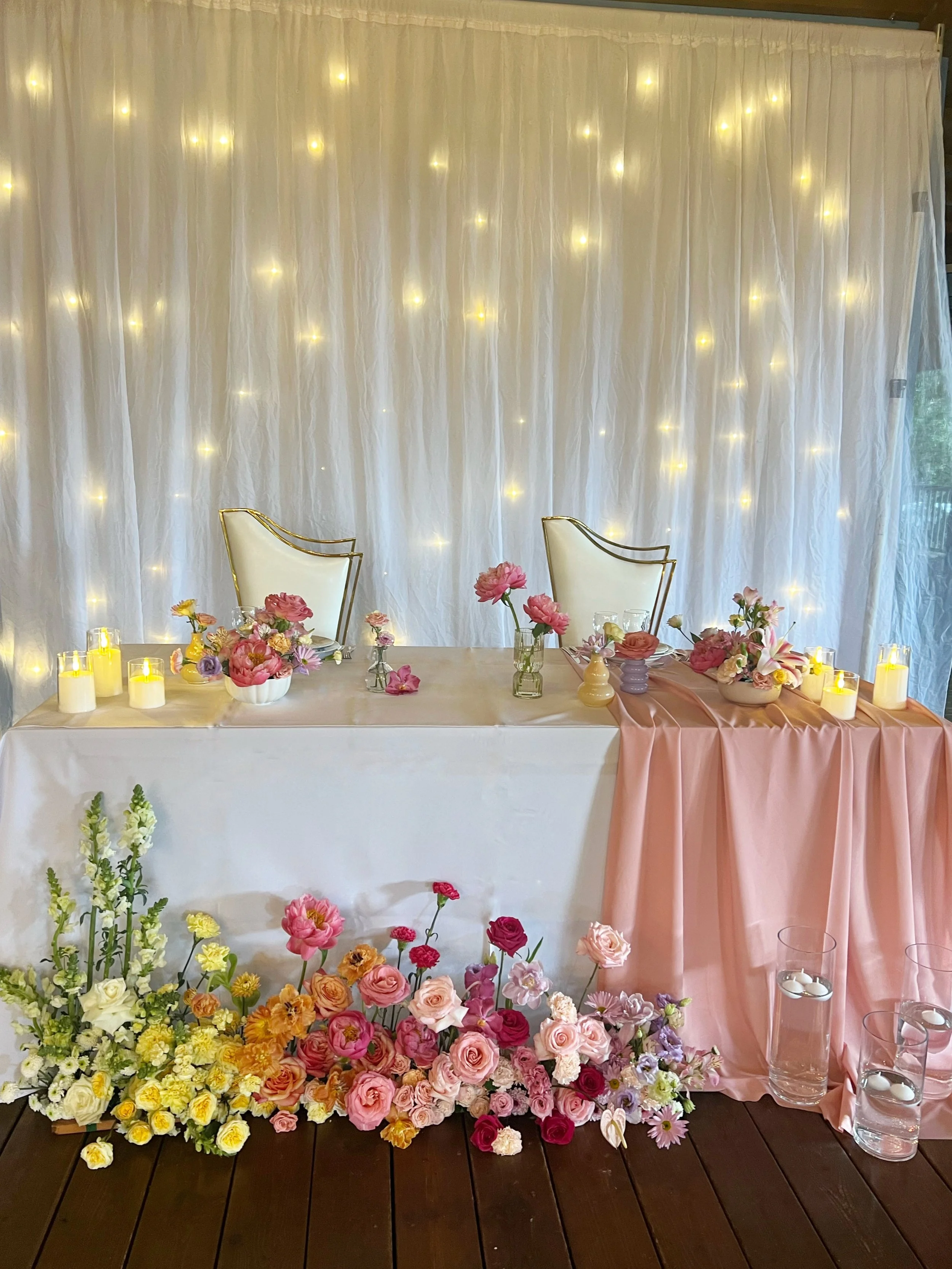 Décor de mariage avec un fond de guirlandes lumineuses derrière une table magnifiquement décorée de fleurs et de bougies.