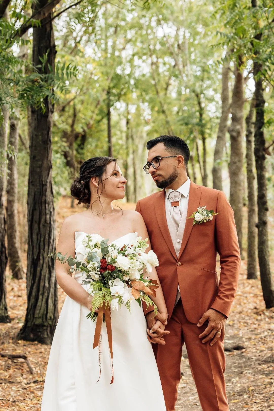 Un couple de mariés, un homme et une femme, se tenant par la main dans une forêt. La mariée porte une robe blanche et tient un bouquet de fleurs, l'homme porte un costume marron avec une boutonnière. Ils se regardent tendrement.