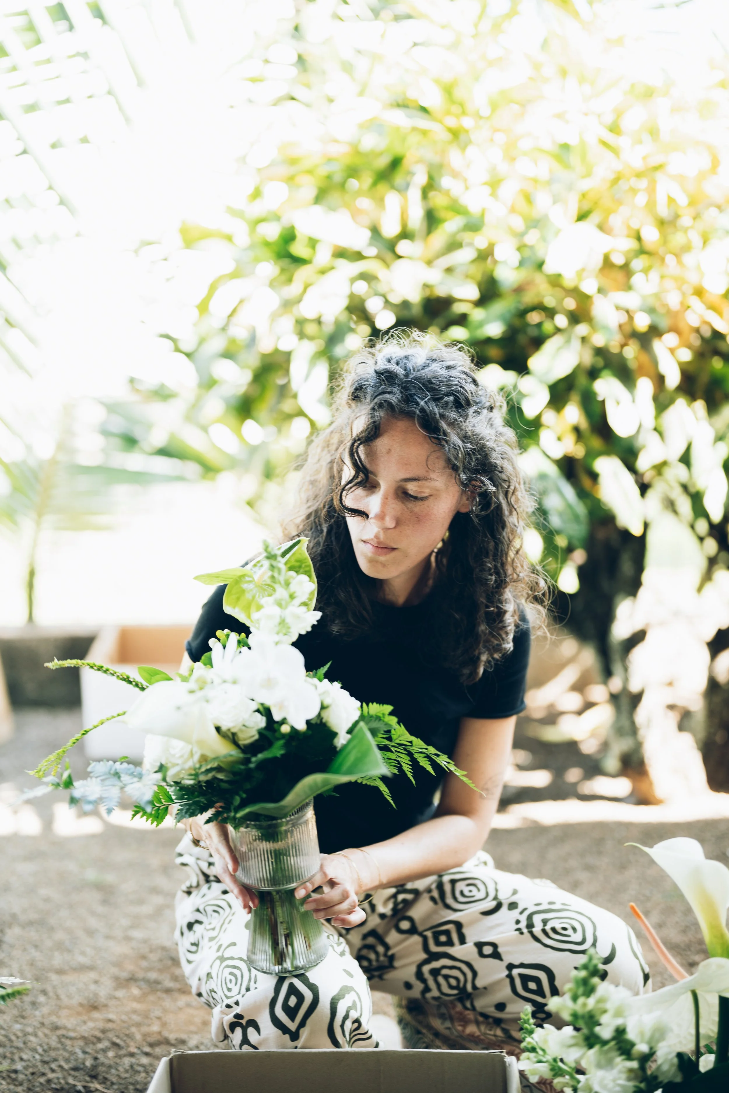 Femme à la peau claire, cheveux longs et bouclés, portant un haut noir et un pantalon à motifs géométriques noirs et blancs, assemble un bouquet de fleurs blanches et vertes dans un vase en verre. Elle est à l'extérieur, entourée de végétation luxuriante et de lumière naturelle.