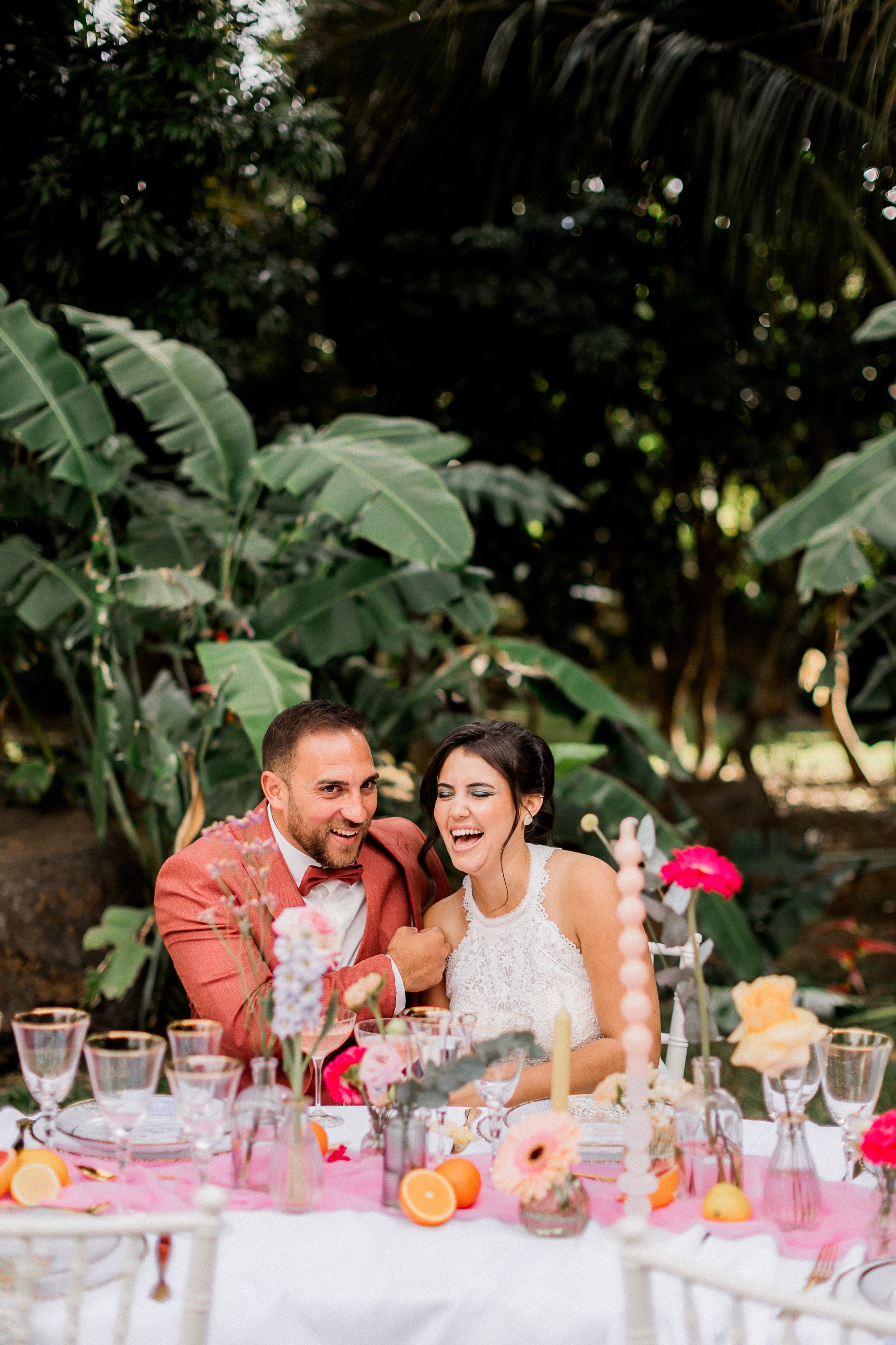 Un couple souriant assis à une table de fête en plein air, décorée de fleurs et de fruits, avec une forêt en arrière-plan.