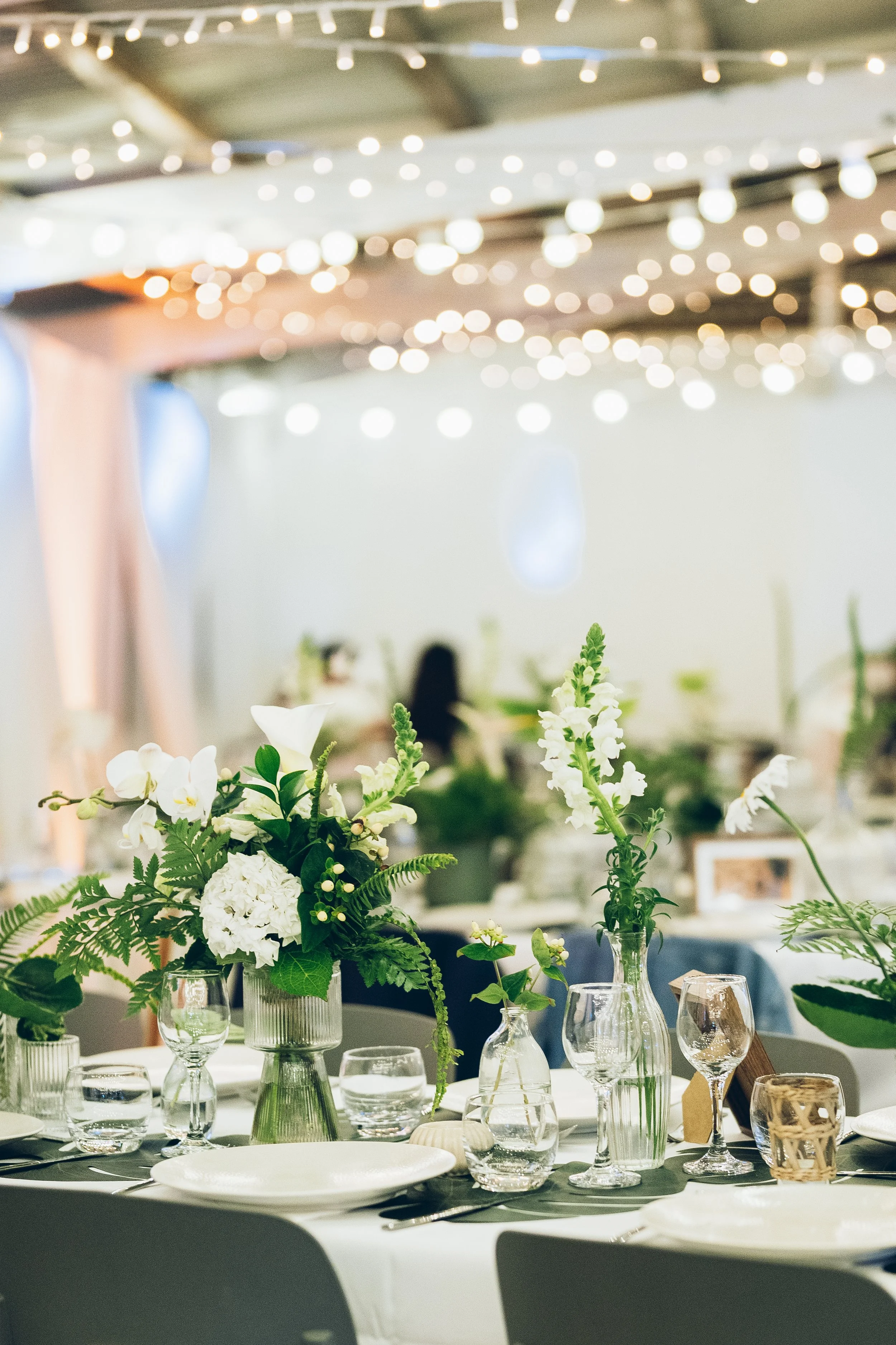 Table décorée avec des fleurs blanches et vertes dans des vases en verre, ambiance élégante avec des guirlandes lumineuses.