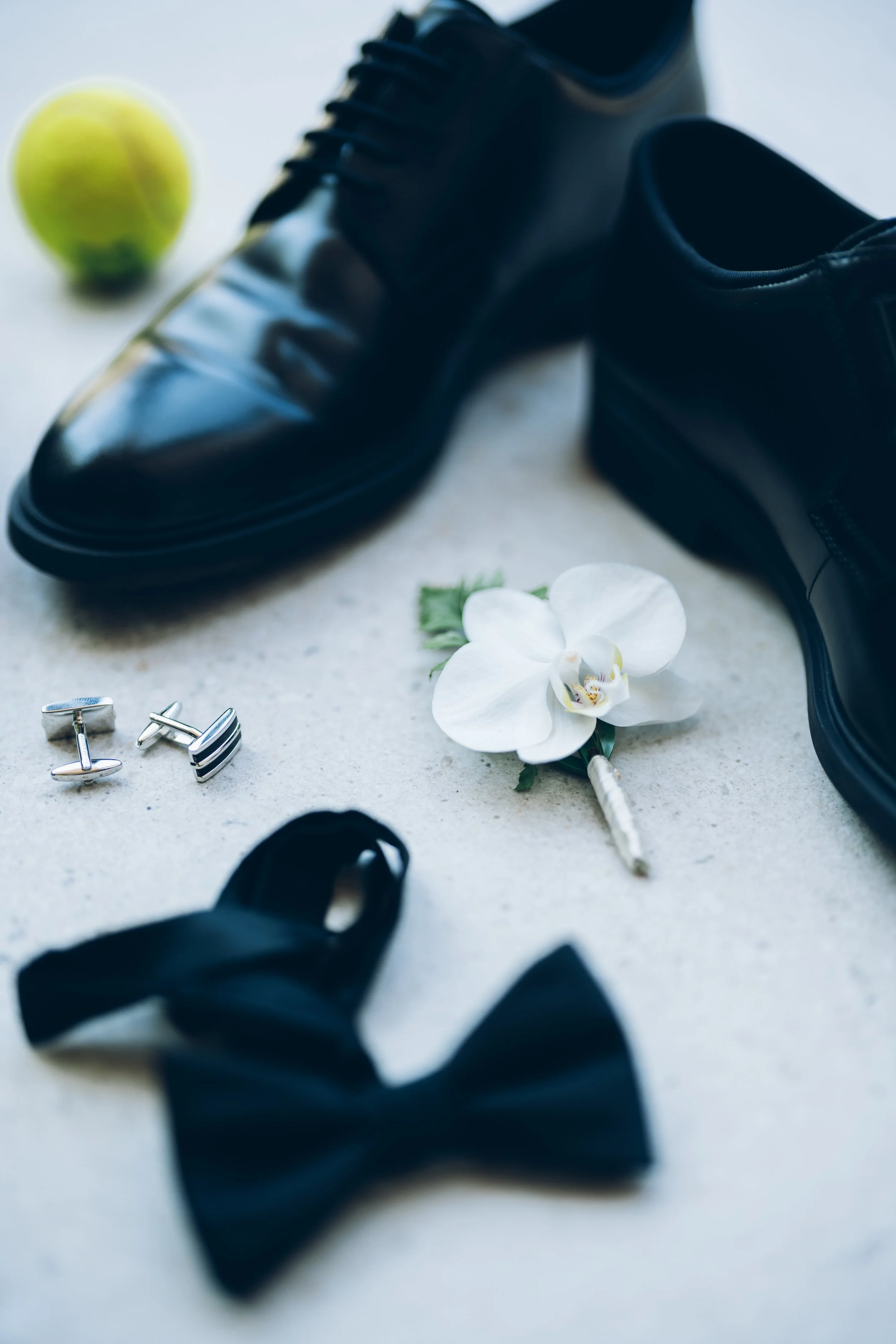 Chaussures de ville en cuir noir, une balle de tennis jaune, des boutons de manchette argentés, une orchidée blanche et un stylo à plume argenté sont disposés sur une surface claire.