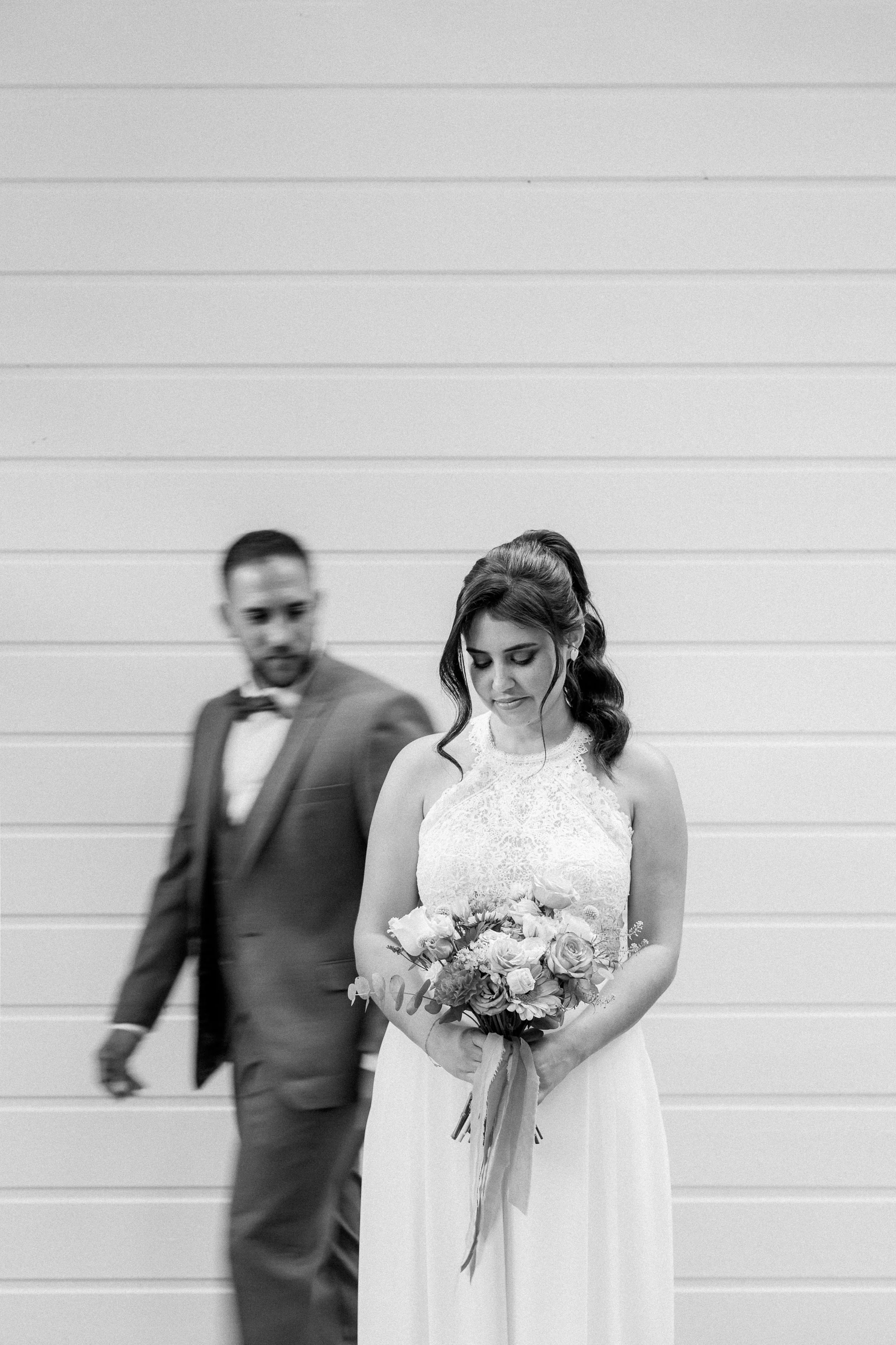 Une femme en robe de mariage tient un bouquet de fleurs, et un homme en costume est flou en arrière-plan.