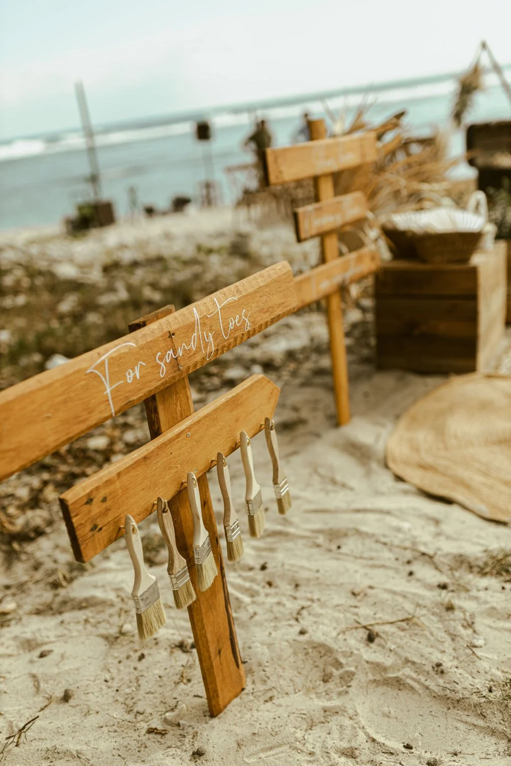 Banc en bois avec un panneau écrit 'For sandy toes' placé sur une plage. Clous et pinceaux attachés au panneau. En arrière-plan, des décorations de plage et la mer.