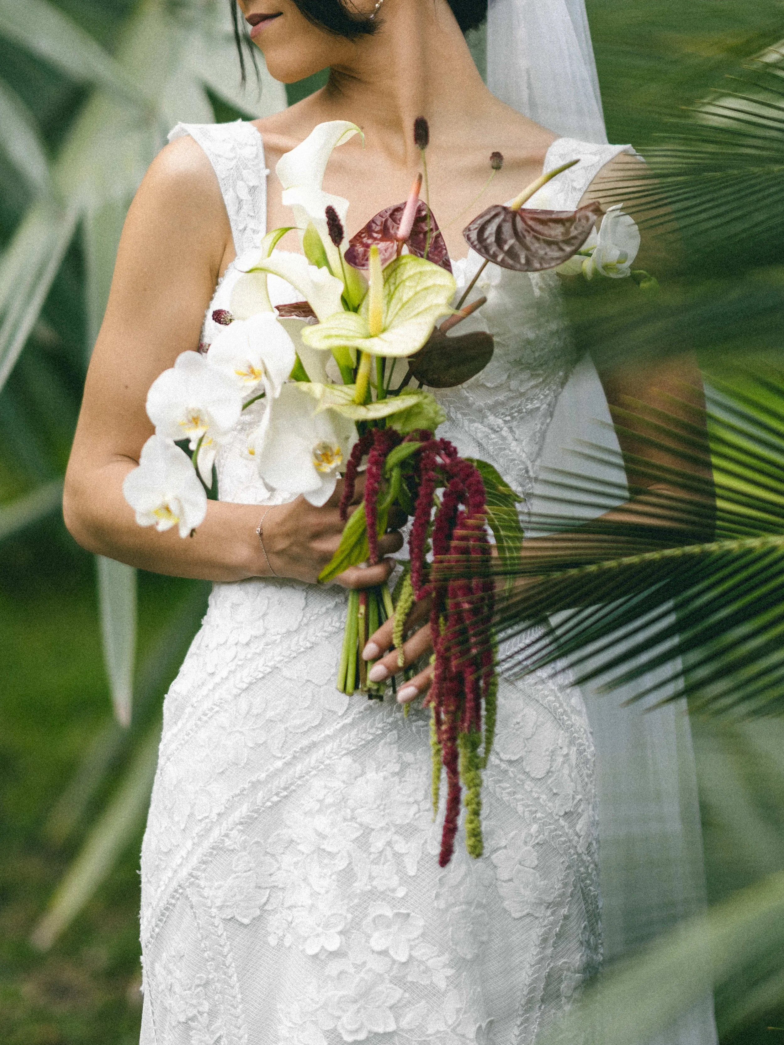 Une femme en robe blanche en dentelle, tenant un bouquet de fleurs composé de callas blanches, d'anthuriums et d'autres fleurs colorées, entourée de feuillage vert.
