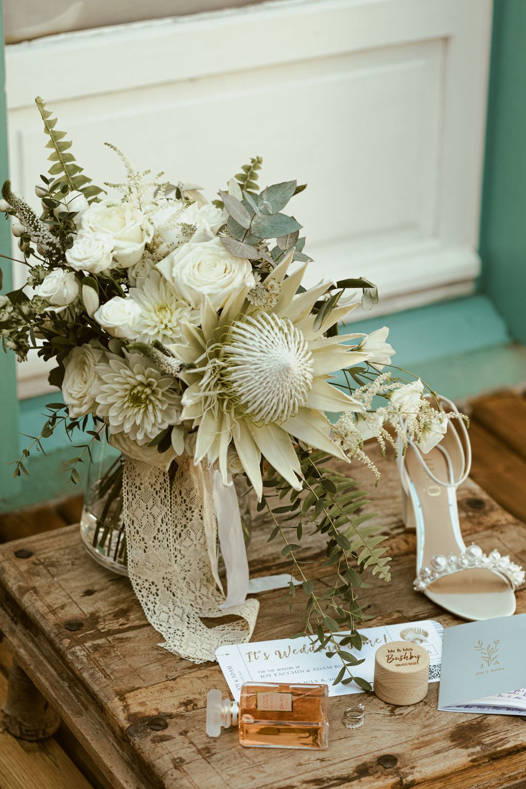 Un bouquet de fleurs blanches avec des roses, des dahlias et d'autres fleurs, dans un vase en verre avec un ruban en dentelle, placé sur une table en bois rustique. À côté, une paire de chaussures de mariage, un parfum en flacon et des accessoires.
