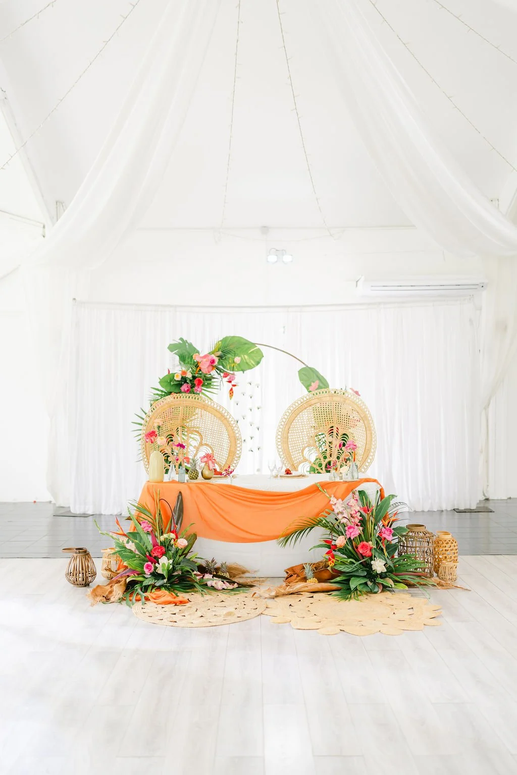 Décoration intérieure avec un thème tropical ou bohème, comprenant deux fauteuils en rotin, un arrangement floral coloré, une table avec un drapé orange, et des accessoires en paille.