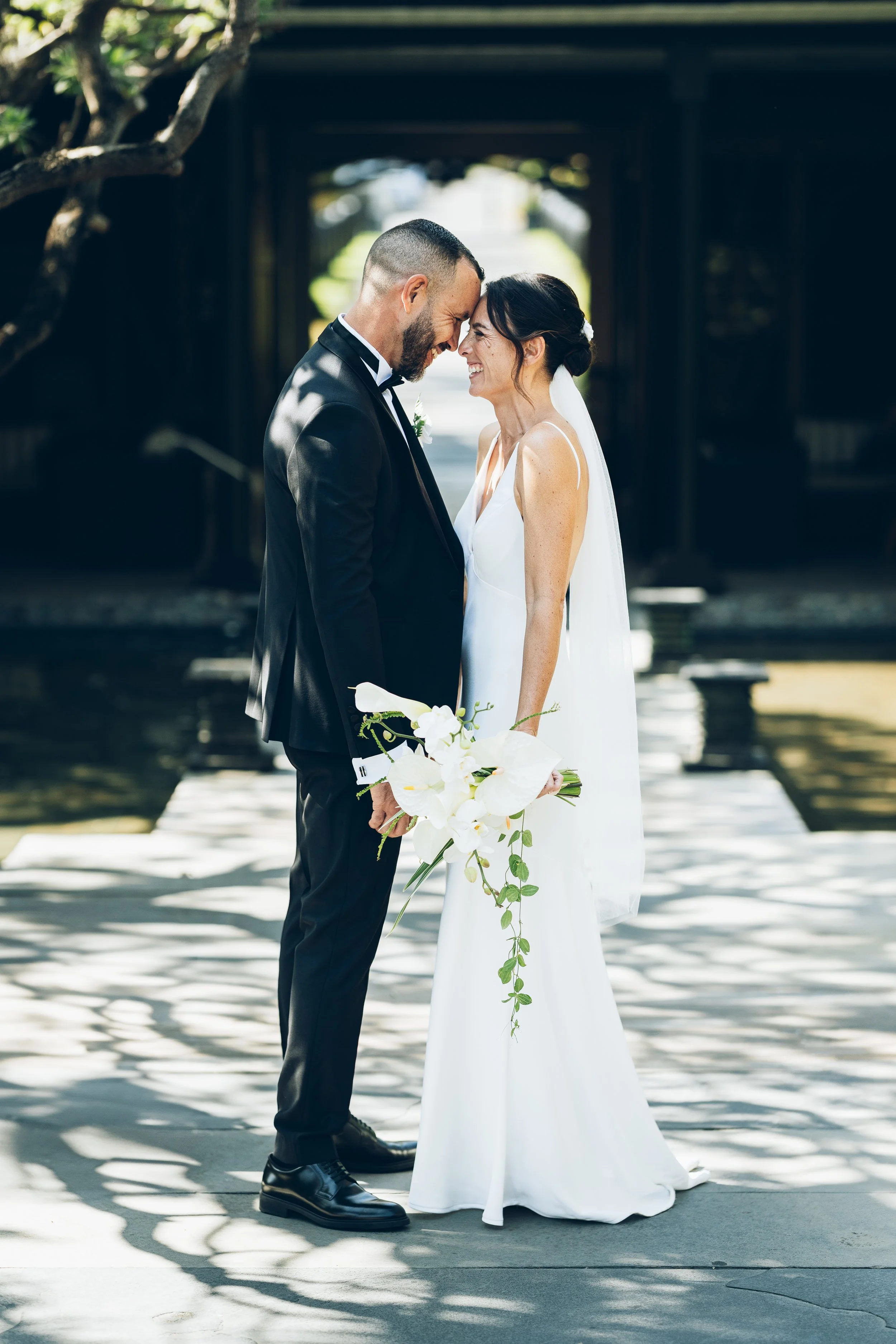Un couple de mariés s'embrassent tendrement devant un fond flou, sous un arbre, lors d'une célébration en plein air. La mariée porte une robe blanche avec un voile et tient un bouquet de fleurs blanches. L'homme porte un costume noir avec une boutonn