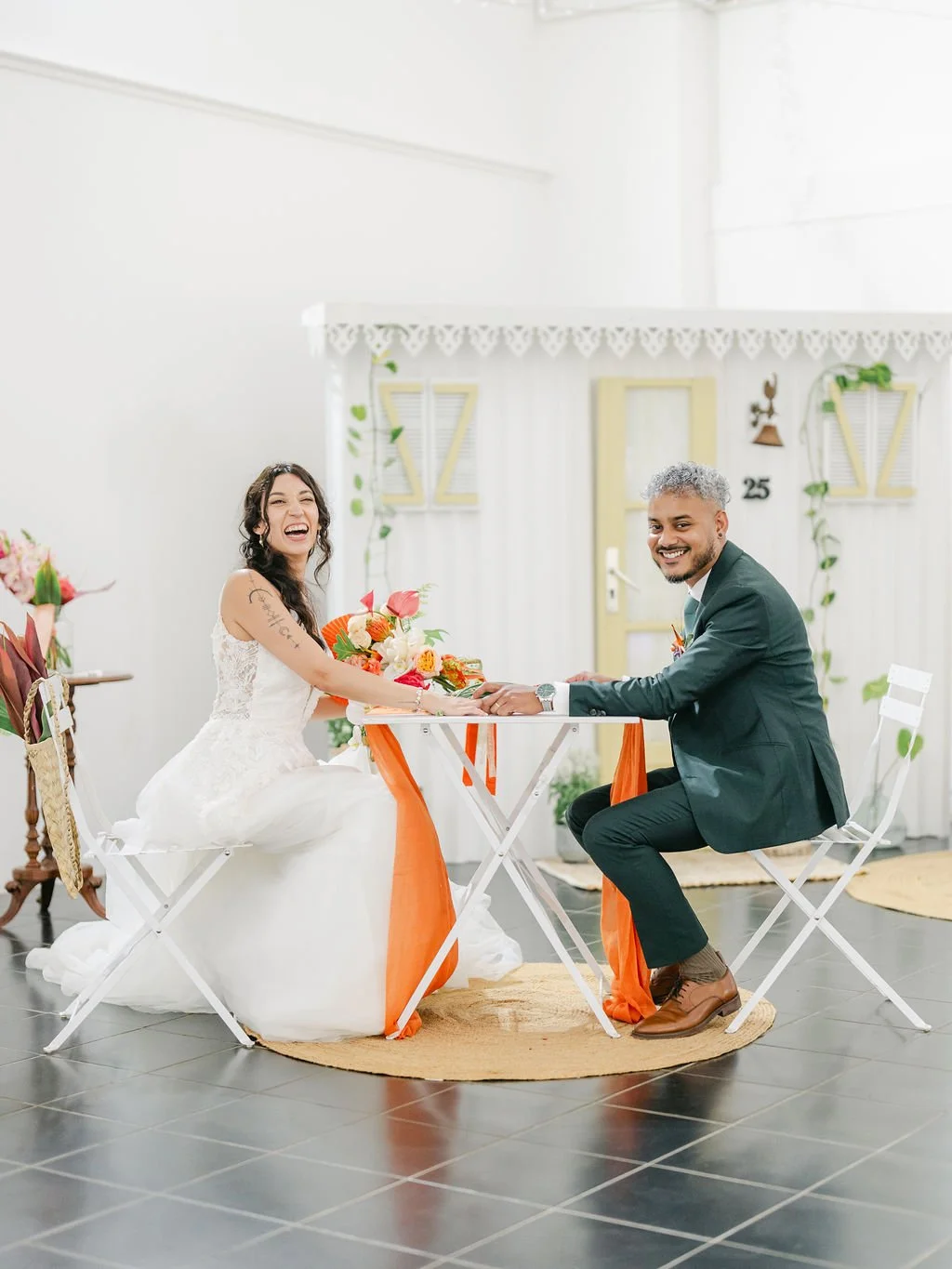 Un couple marié lors de leur cérémonie de mariage, assis à une petite table avec un bouquet de fleurs, dans une salle décorée avec des éléments colorés et une atmosphère joyeuse.