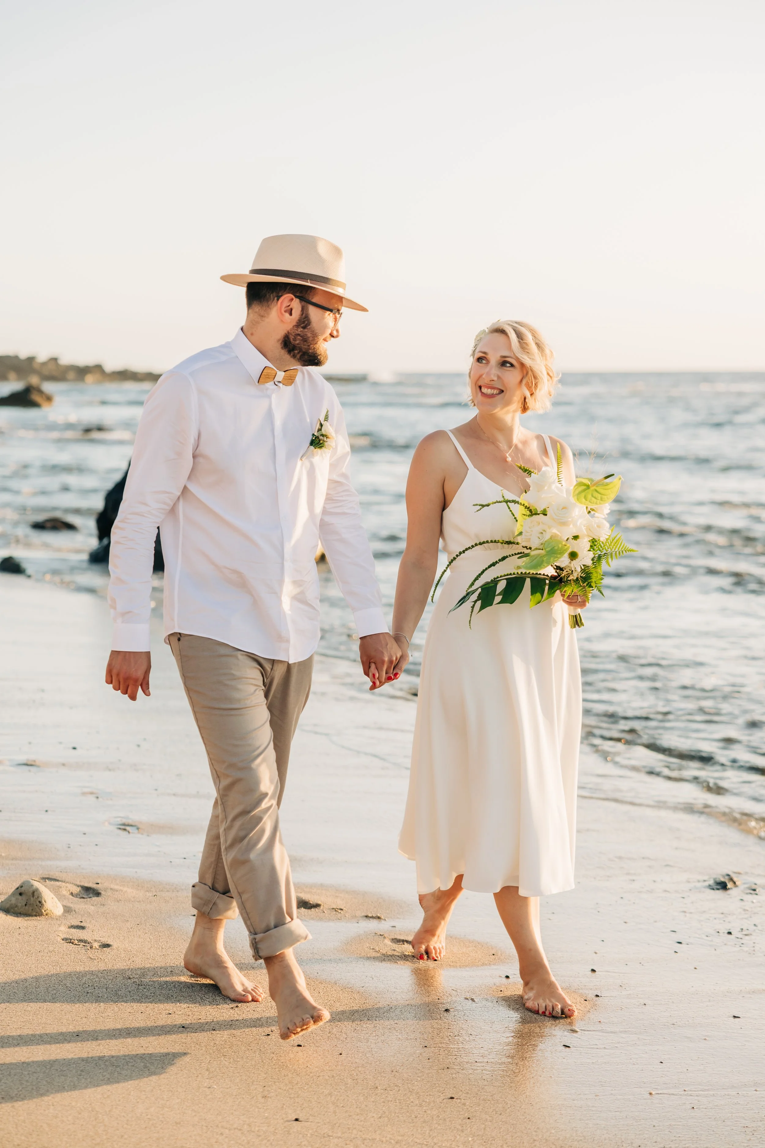 Un couple marchant main dans la main sur la plage, en surfeur de vêtements décontractés, avec la mer en arrière-plan, sable et rochers.