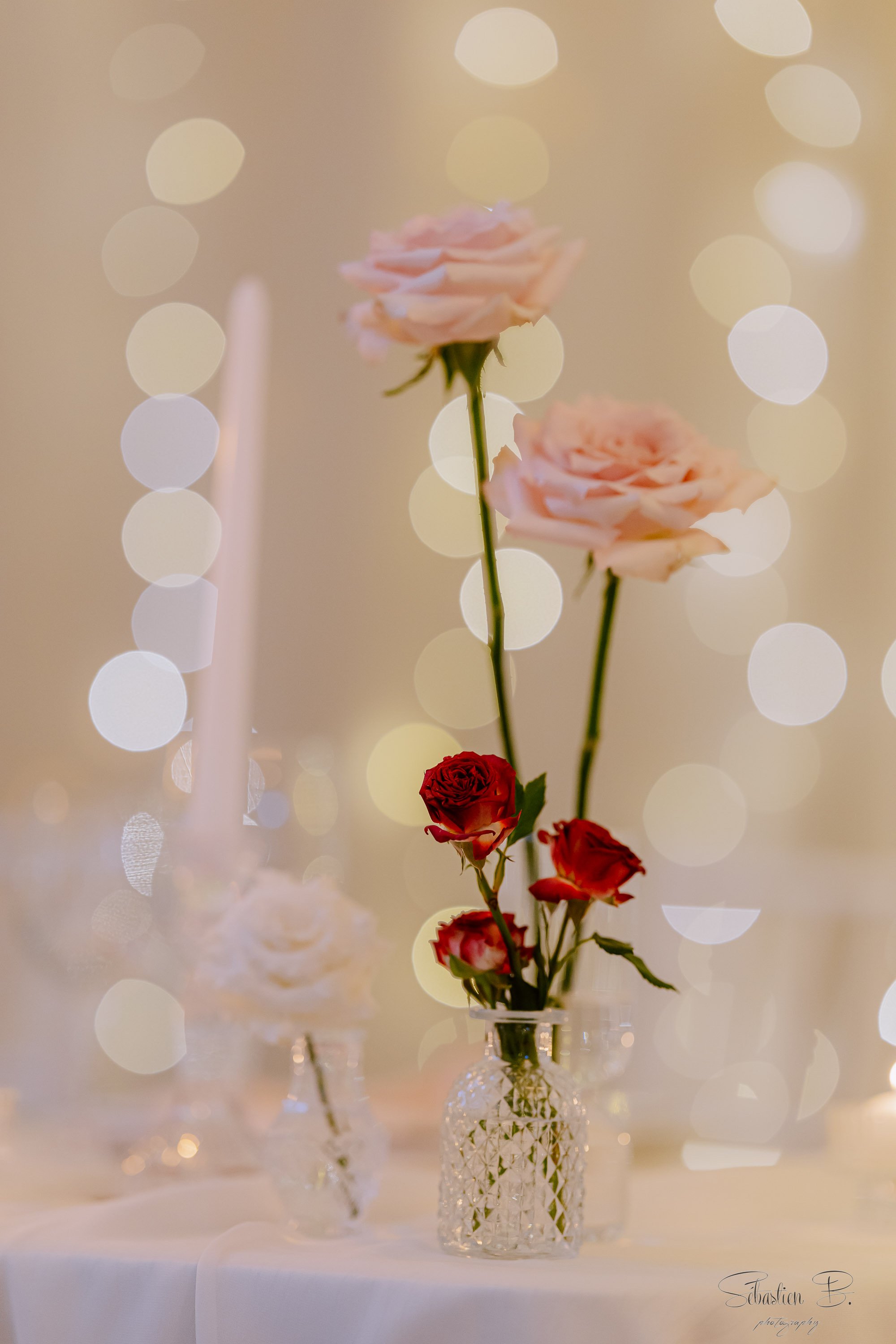 Un bouquet de roses pale en vase en verre, avec des roses rouges en dessous, sur une table blanche avec un fond de lumières bokeh blanches et crème.