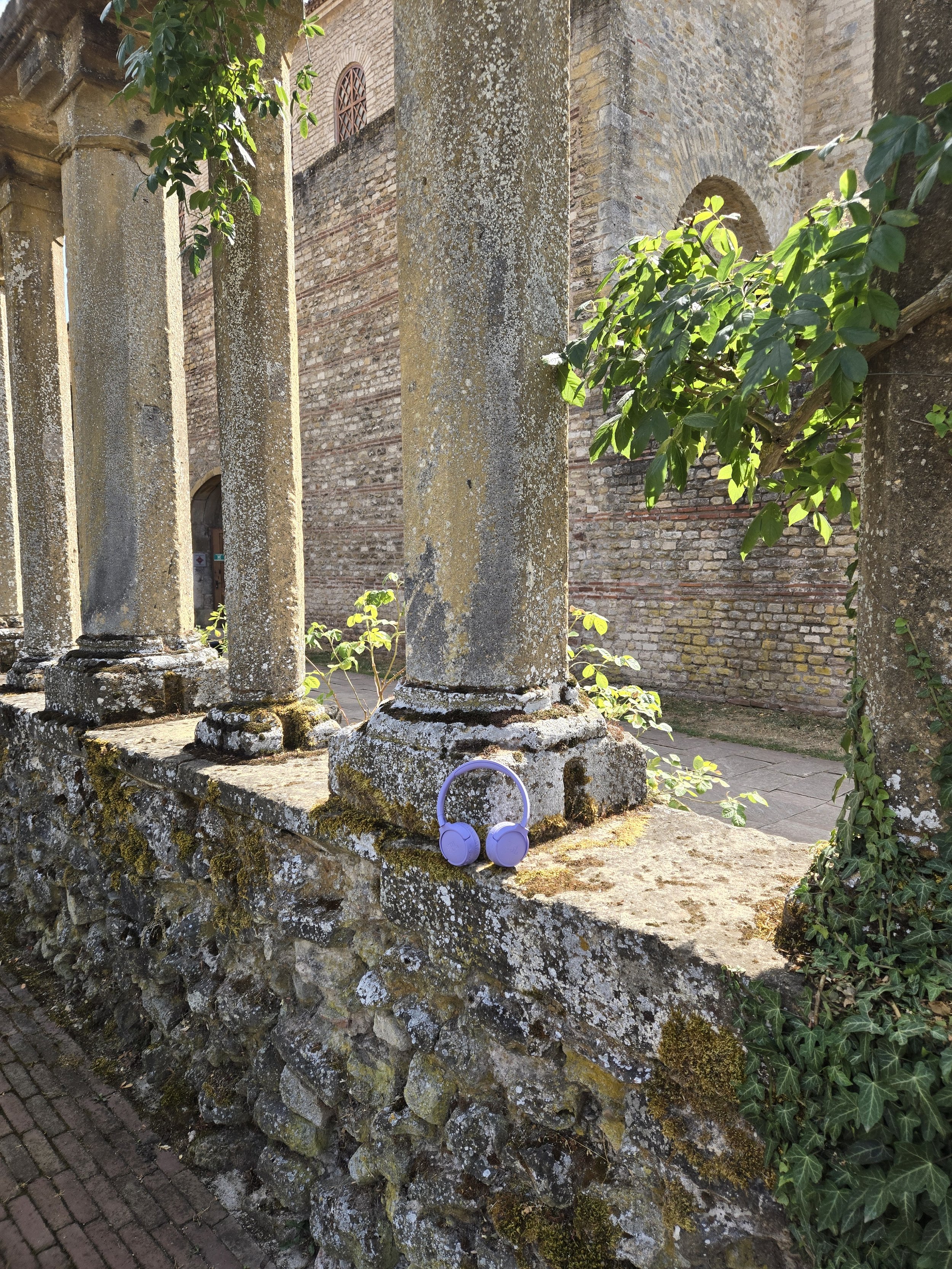 Des anciennes colonnes en pierre recouvertes de mousse, situées à côté d'un mur en briques, avec une paire de casque audio violets suspendus à la pierre au premier plan. La scène est illuminée par la lumière naturelle et des végétaux verts, notamment de la vigne grimpante.