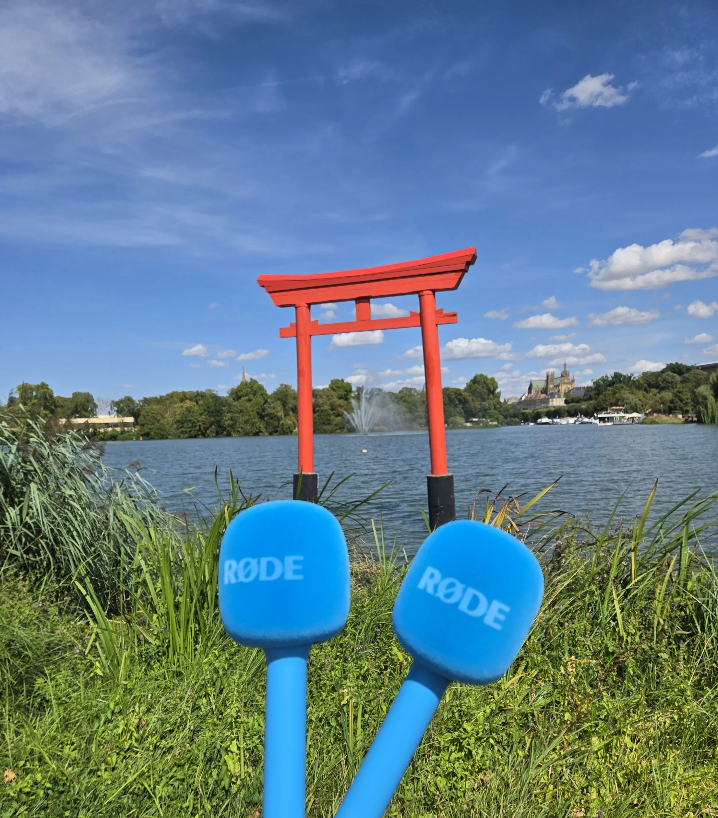 Photo d'un lac avec une fontaine, en arrière-plan un château et un ciel bleu avec quelques nuages, vue partielle de deux ballons bleus portant le mot "RODE" sur l'un d'eux, et une structure rouge en forme de torii japonais.