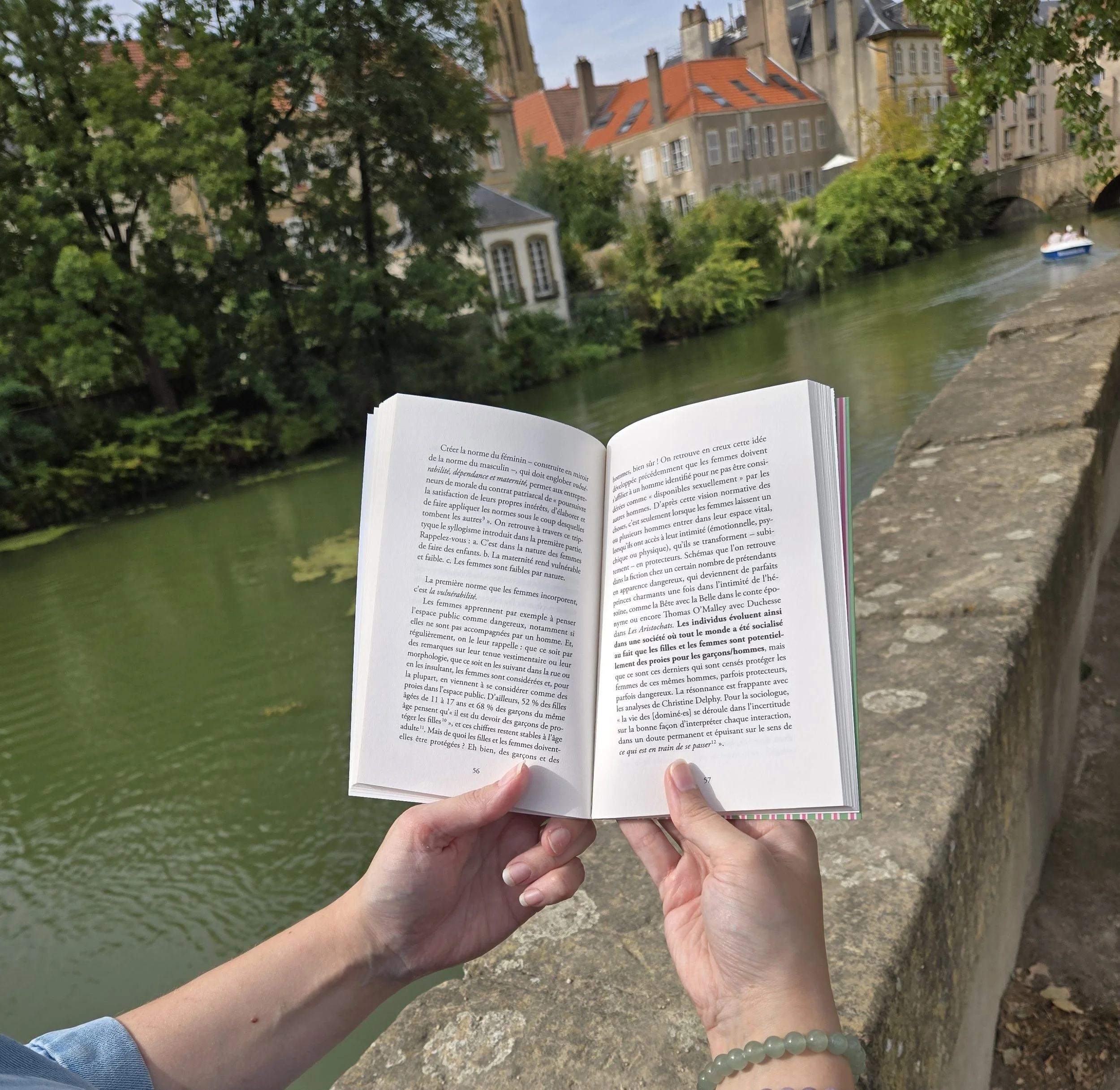 Une personne tient un livre ouvert avec un texte en français, près d'une rivière bordée d'arbres et de bâtiments historiques en pierre, avec un petit bateau à moteur sur l'eau, en extérieur par une journée ensoleillée.