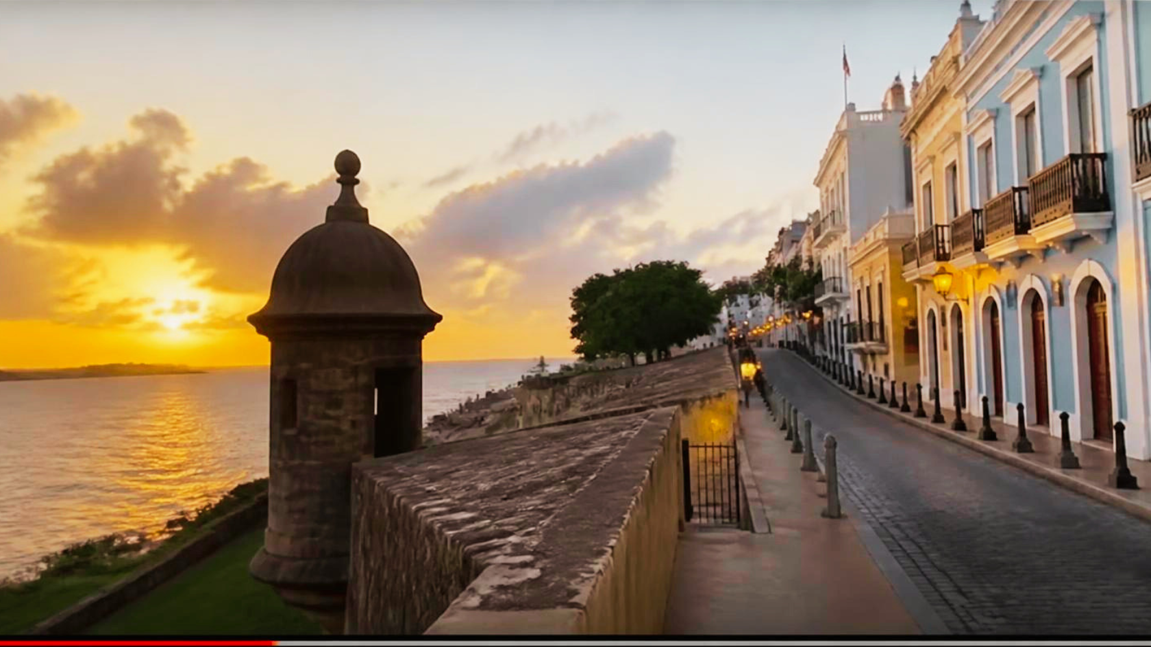 Sunset over a waterfront street with colorful historic buildings, a stone fortification wall, and trees along the promenade.