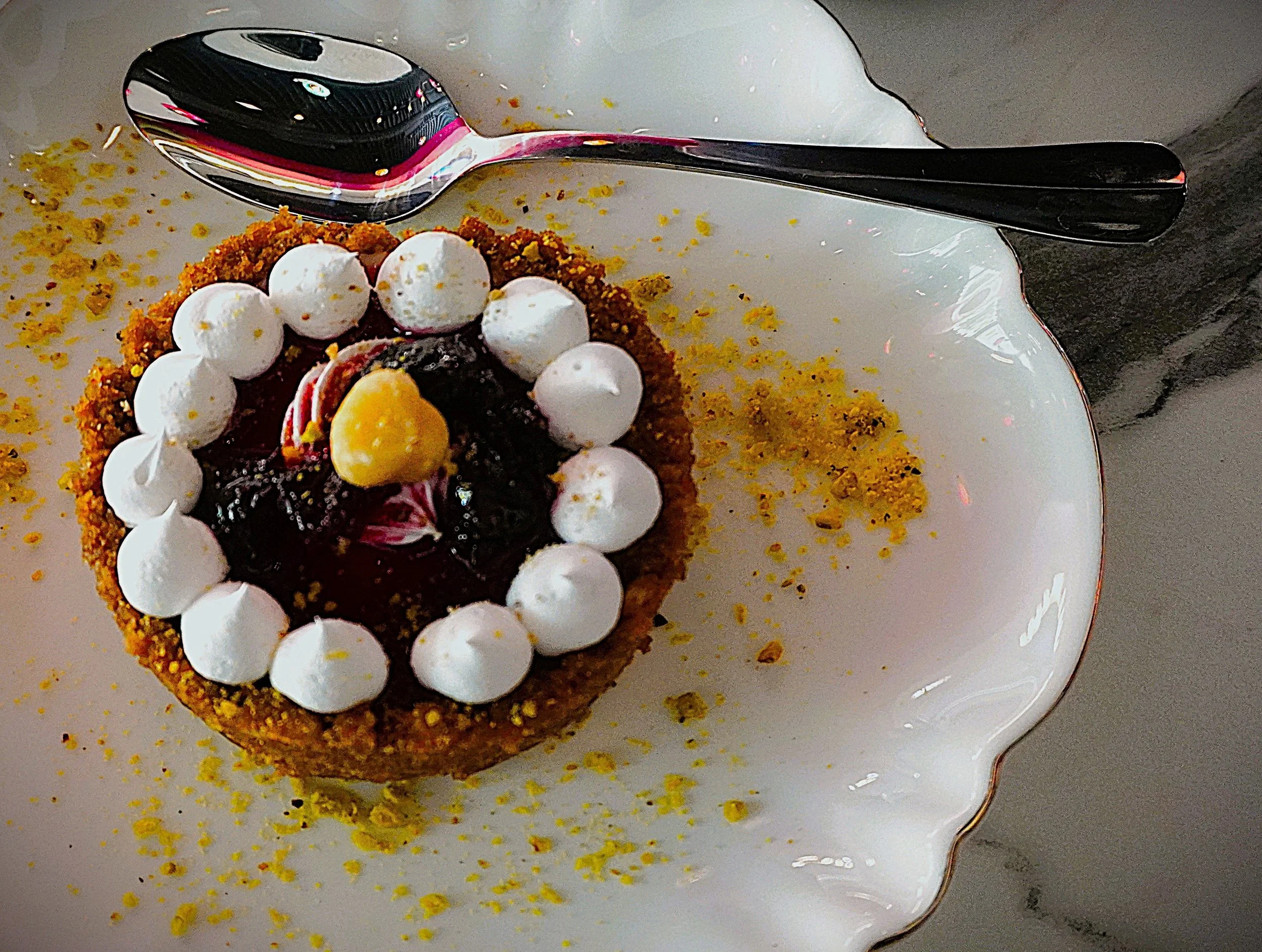 A partially visible dessert on a white plate, decorated with dollops of whipped cream, a yellow flower-shaped decoration, berry topping, and yellow crumbs, with a silver spoon on the plate.