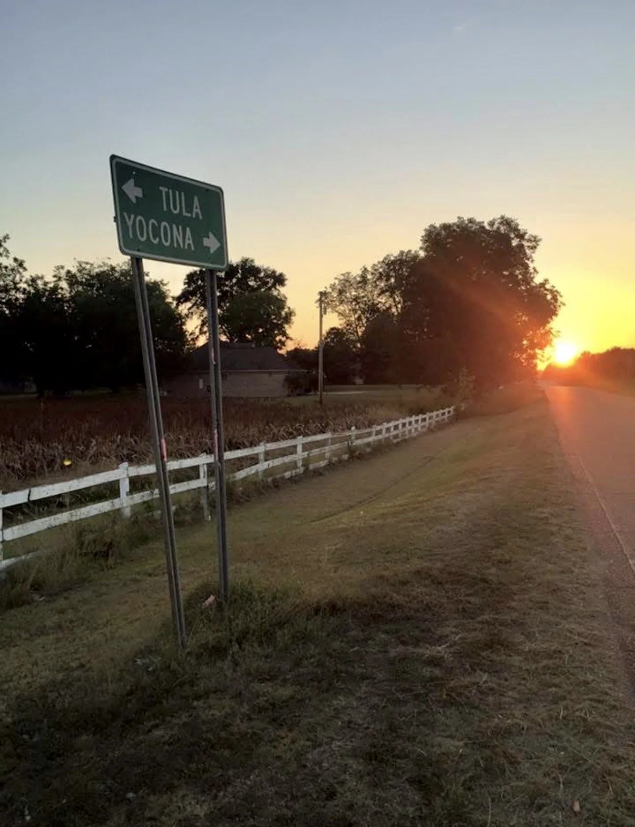 Roadside scene at sunset with a green directional sign indicating left for Tula and right for Yocona. There is a white fence on the left, trees in the background, and the sky is clear with the sun near the horizon.
