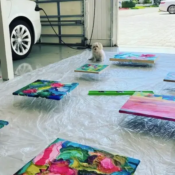 A small dog sitting on a plastic sheet in a garage surrounded by colorful abstract paintings on canvas.