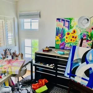 Home art studio with a colorful abstract painting, a black desk, a roll of paper, and art supplies on a table.