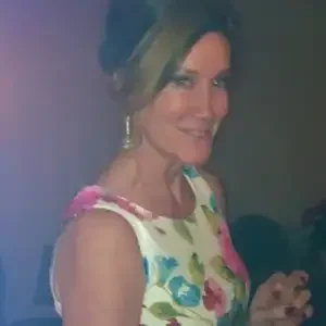 A woman with dark hair tied up, wearing a floral dress and earrings, smiling at the camera.