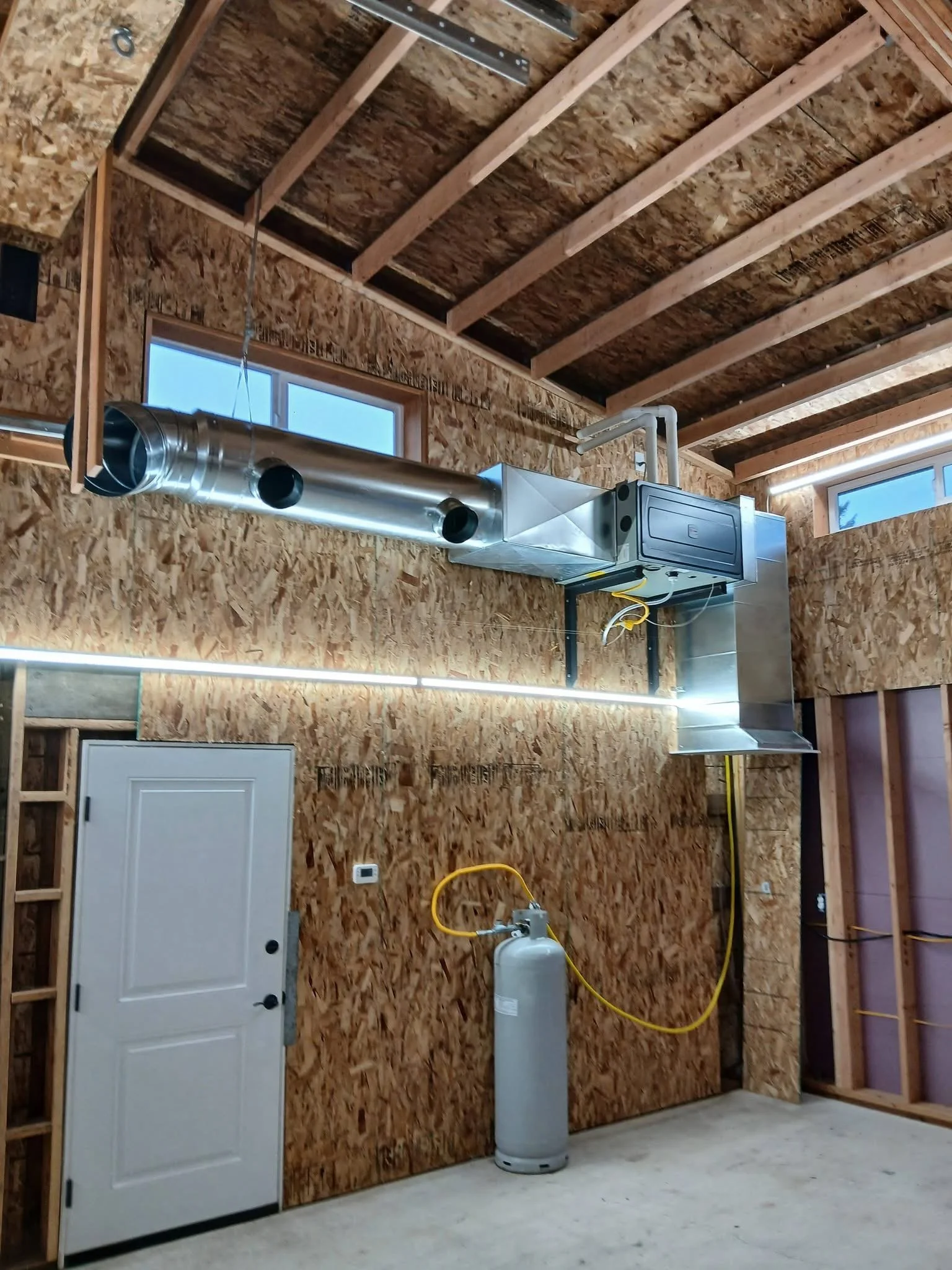 An unfinished garage interior with wood wall panels, exposed ceiling beams, a vent pipe, an electrical panel, and a tank.