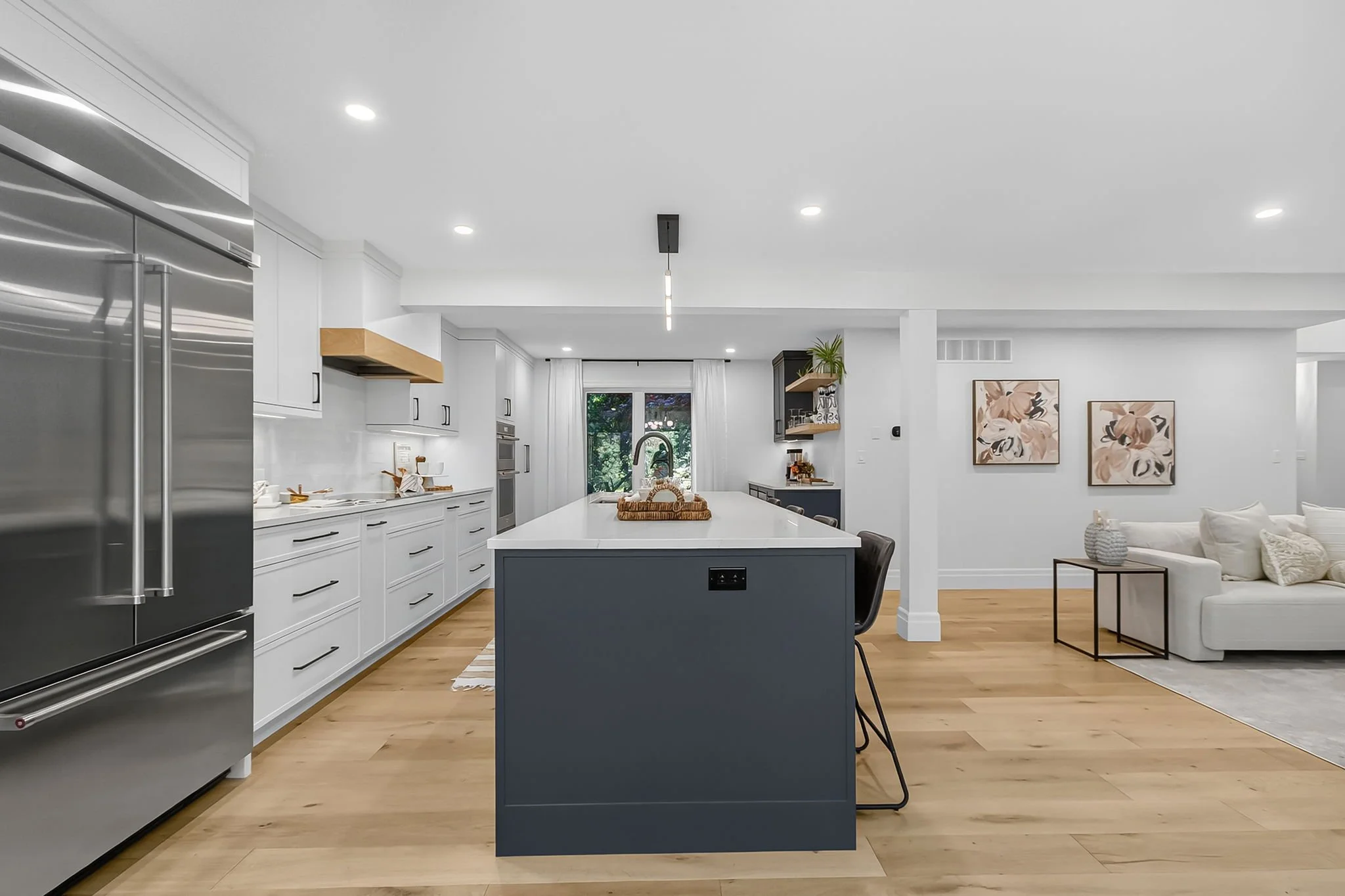Modern kitchen with white cabinetry, stainless steel appliances, a blue central island, and a light wood floor, adjacent to a living area with a white sofa and wall art.