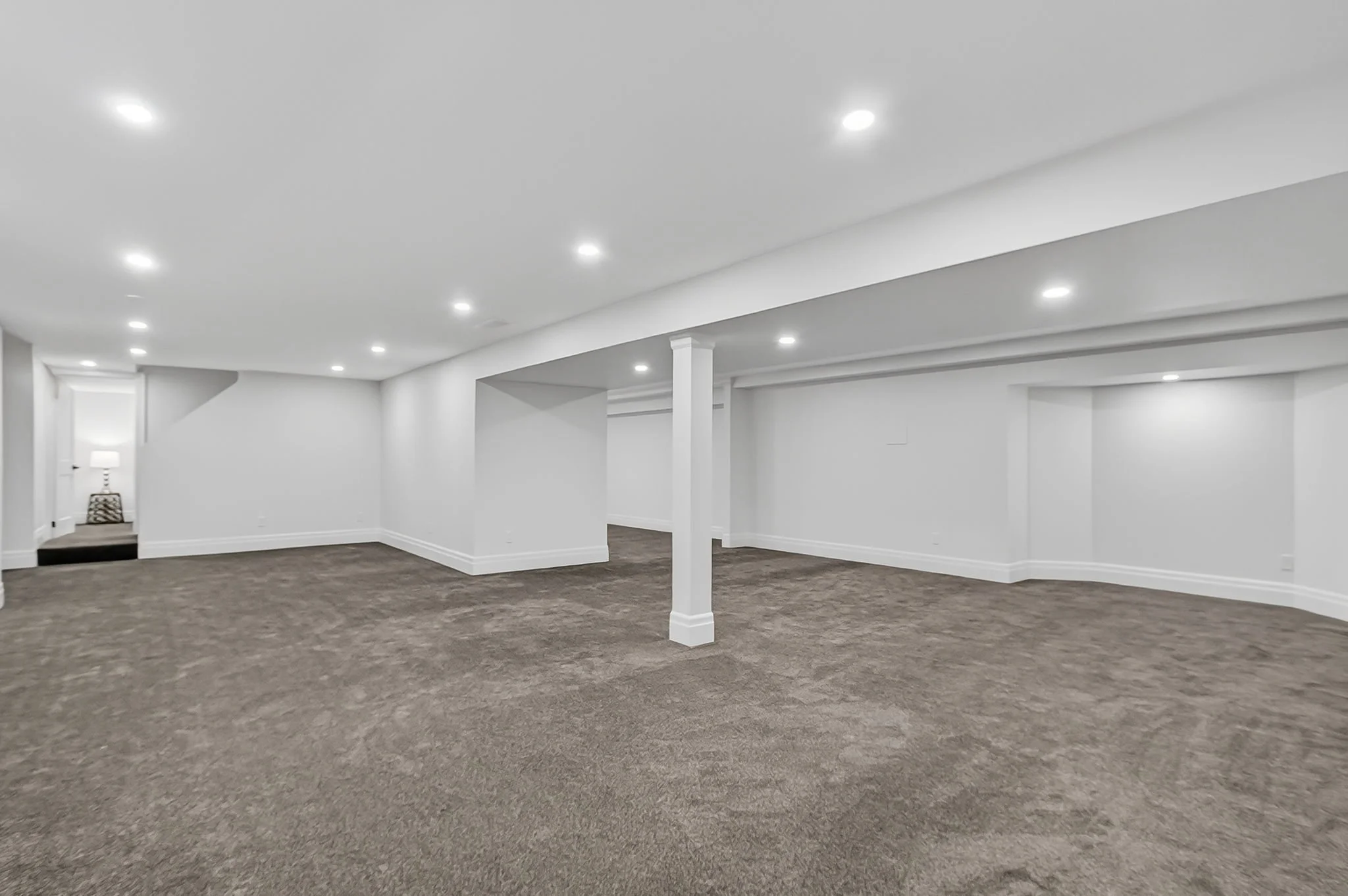 Empty basement room with white walls, ceiling, and ceiling lights, with brown carpet flooring and a corner with a side table and lamp.