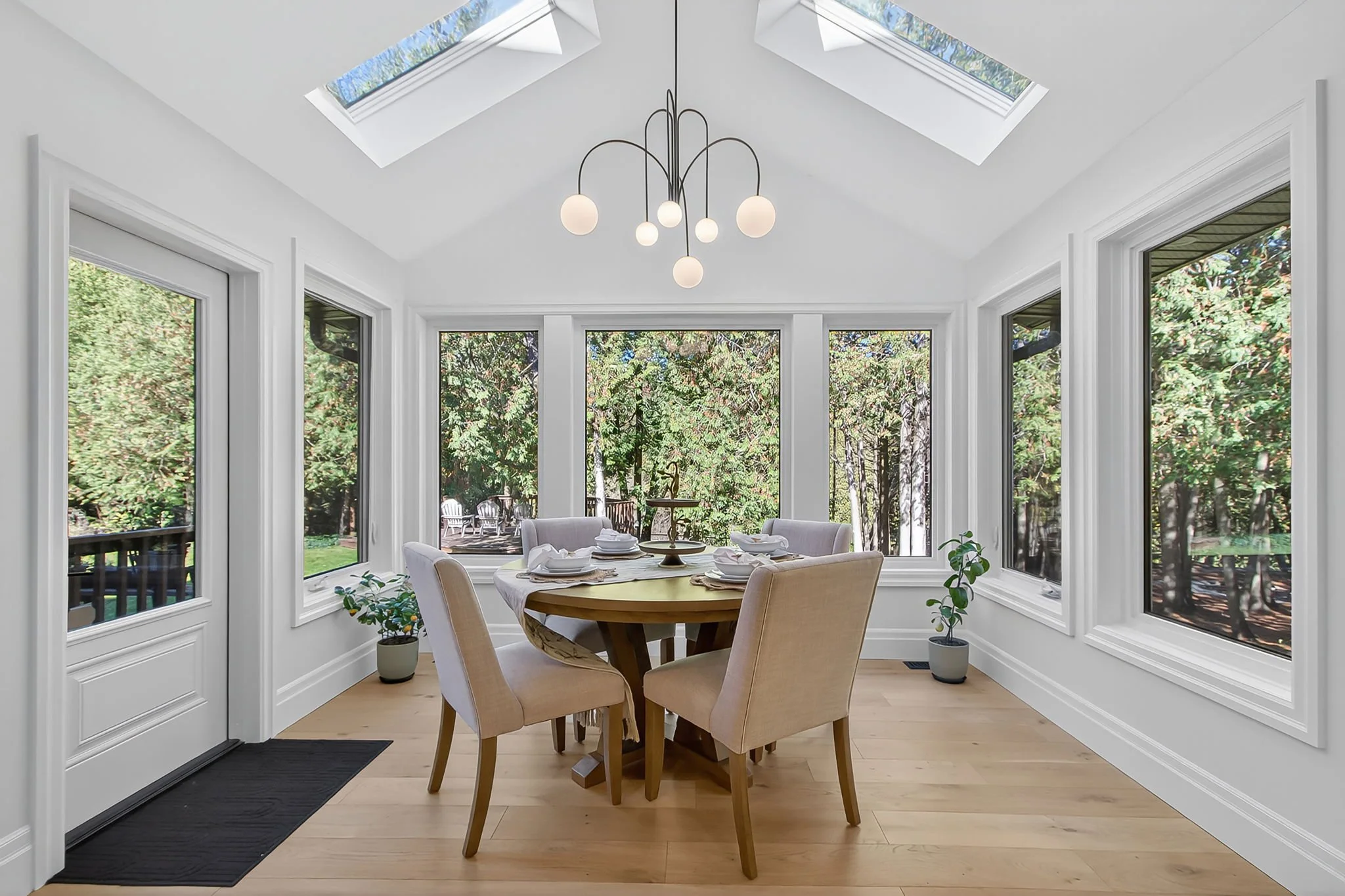 Bright dining room with skylights, large windows, and a round wooden table set for a meal with six beige upholstered chairs. Potted plants are on either side, and a modern chandelier hangs from the ceiling.
