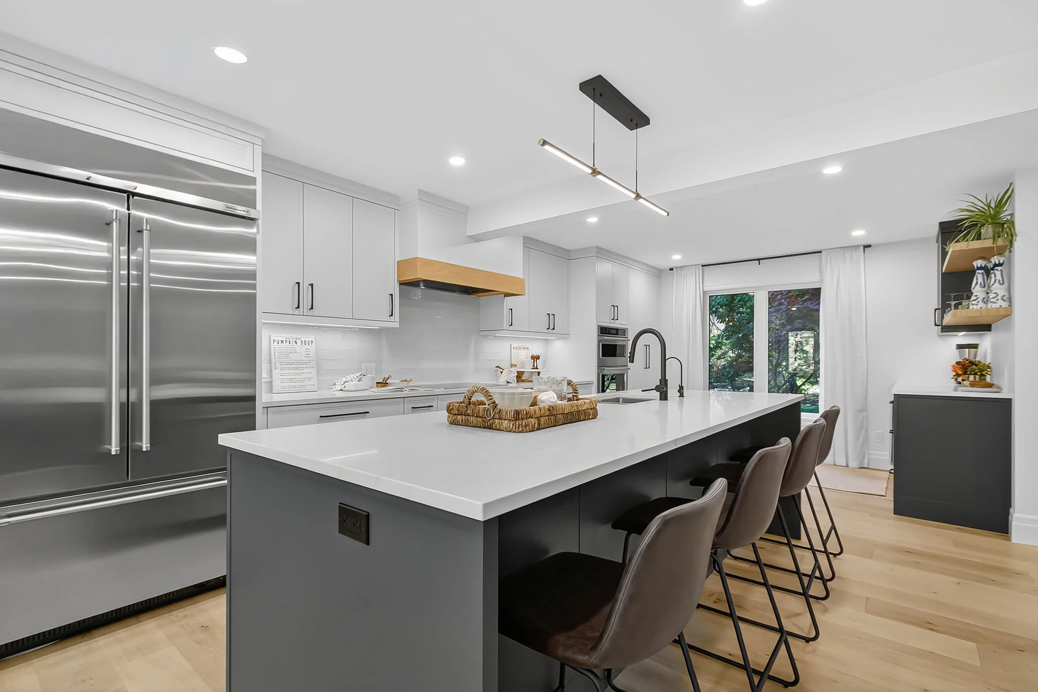 Modern kitchen with white cabinets, stainless steel refrigerator, and an island with seating. There is a hanging light fixture, large windows, and decorative shelves with vases and plants.