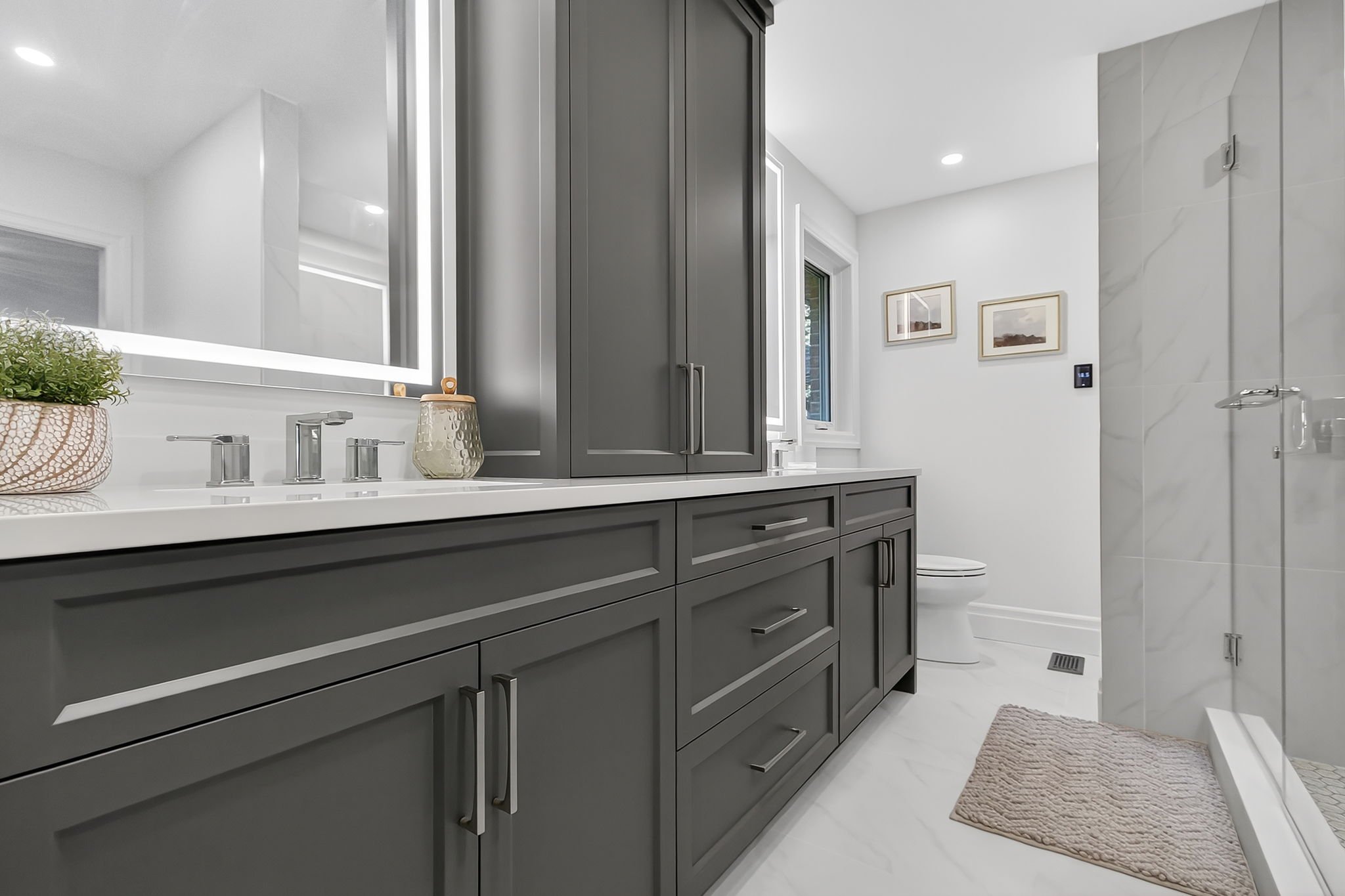 Modern bathroom with gray cabinetry, white countertops, a large mirror with LED lighting, framed art on the wall, and a walk-in shower with a marble wall and a beige bath mat.