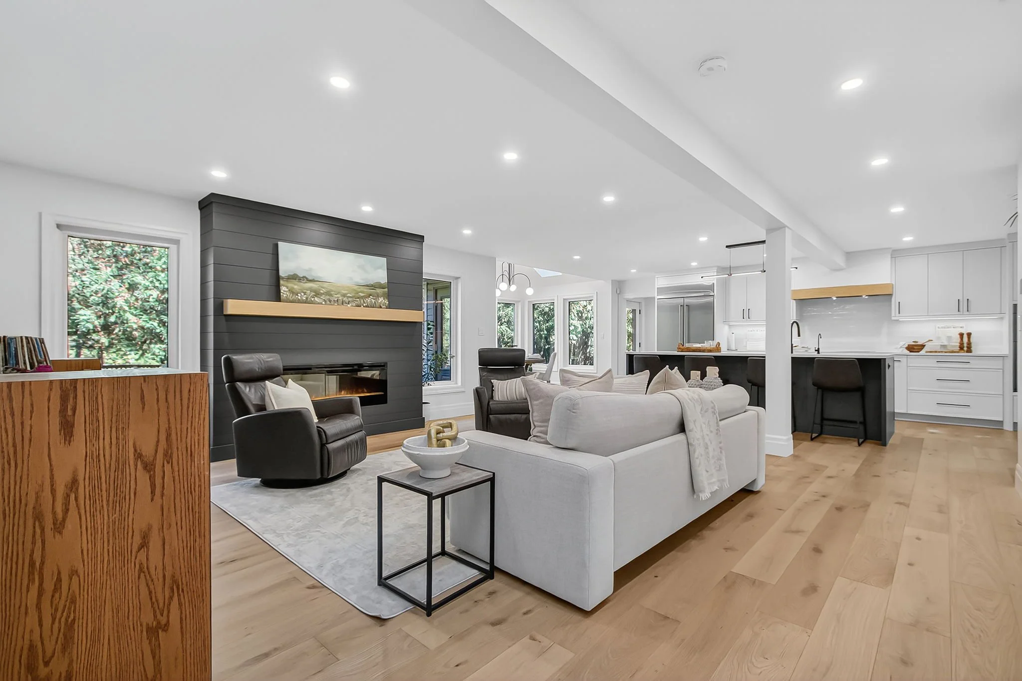 Open-concept living room and kitchen with white walls, hardwood floors, large windows, a gray accent wall with a fireplace, and modern furniture including a white couch, black chairs, and a round side table.