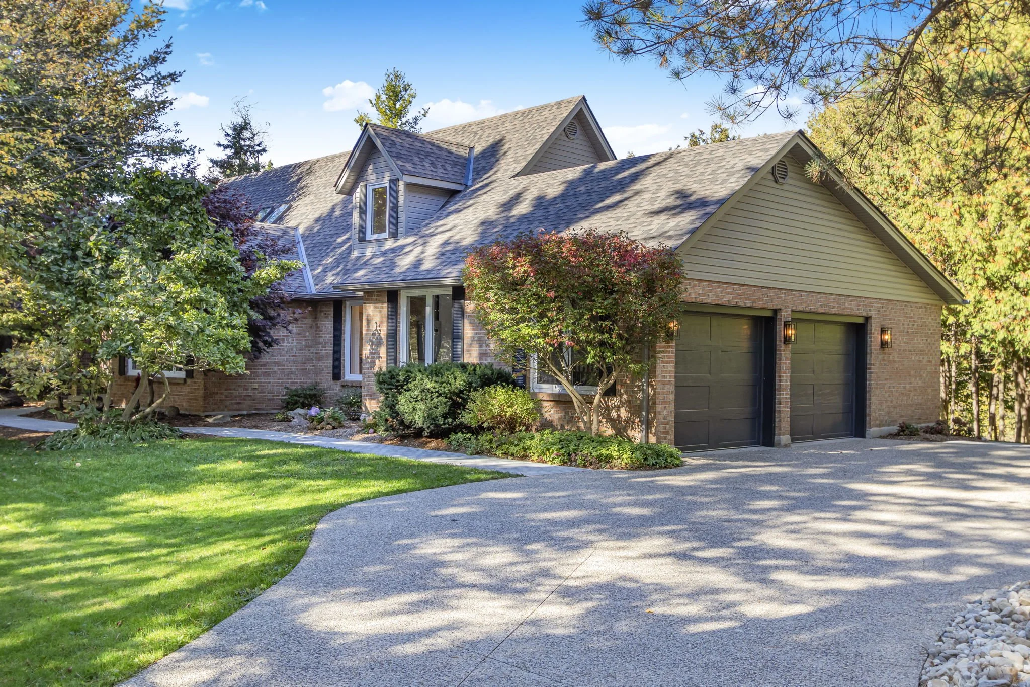 A two-story house with a brick and siding exterior, surrounded by trees and greenery, with a curved concrete driveway leading to two garage doors.