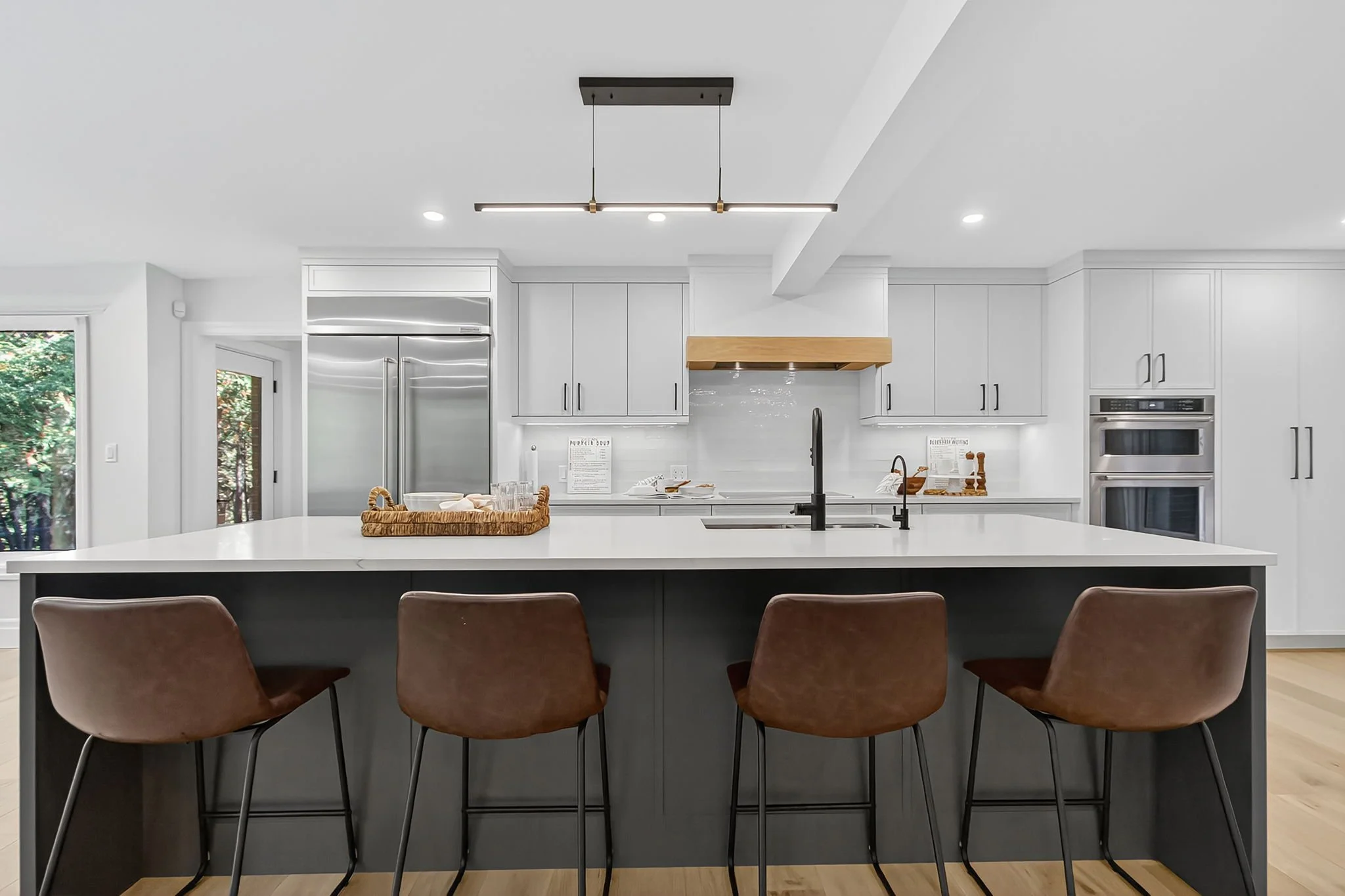 Modern kitchen with white cabinets, stainless steel appliances, a large island with a white countertop, and brown chairs in the foreground.