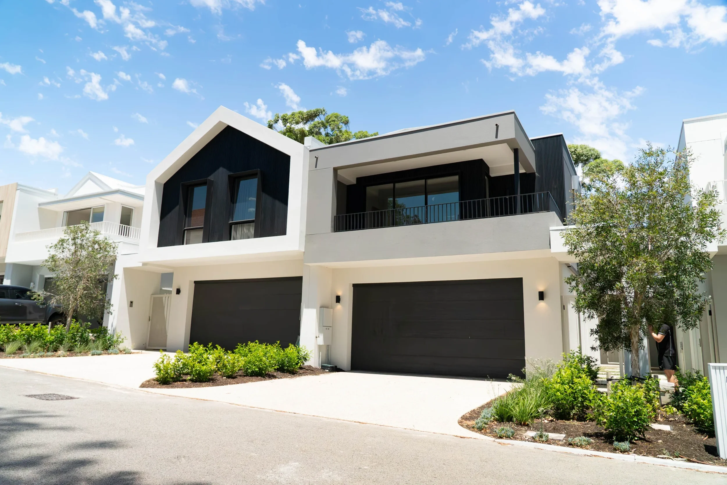 Modern multi-story house with black and white exterior, large black garage doors, balcony, surrounded by trees and greenery under a blue sky with scattered clouds.