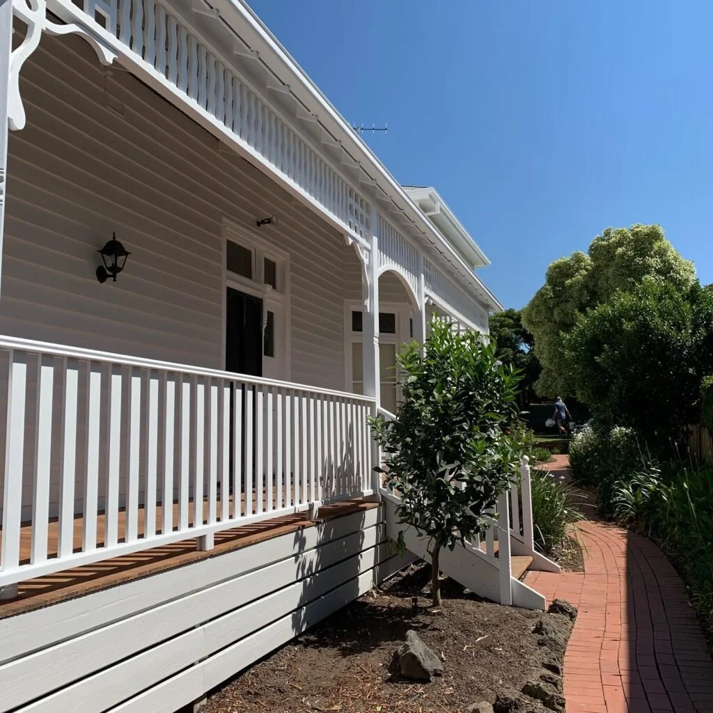A white house with a porch, railing, and stairs leading down to a winding brick pathway surrounded by greenery and trees under a clear blue sky.