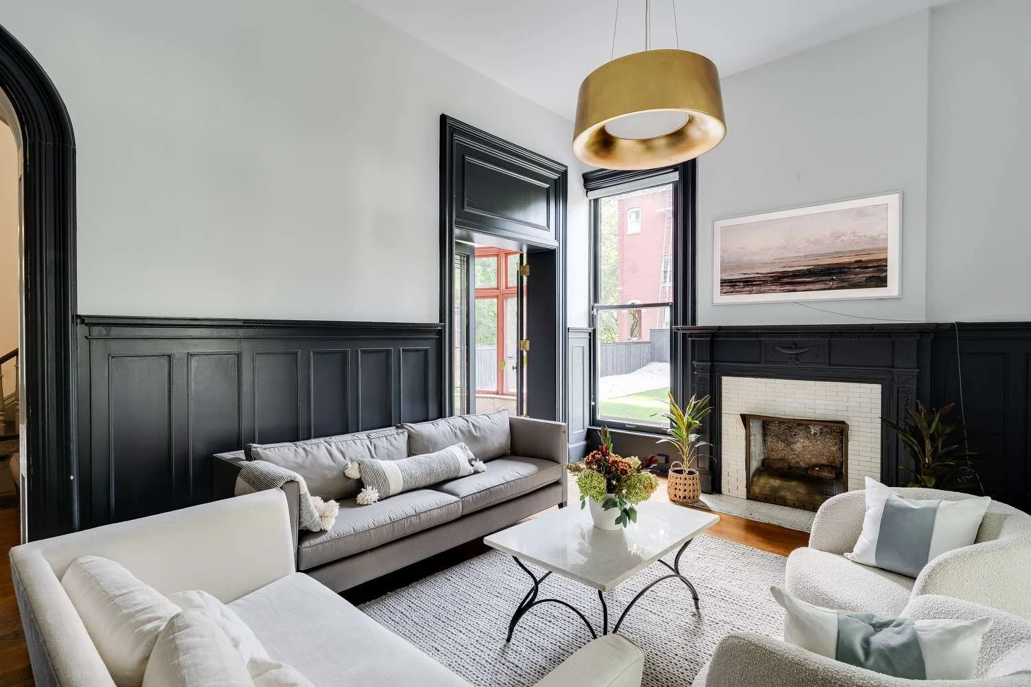 Living room with black-paneled walls, white ceiling, fireplace, floral artwork, beige sofas, a white coffee table with a flower arrangement, large windows, and plants.