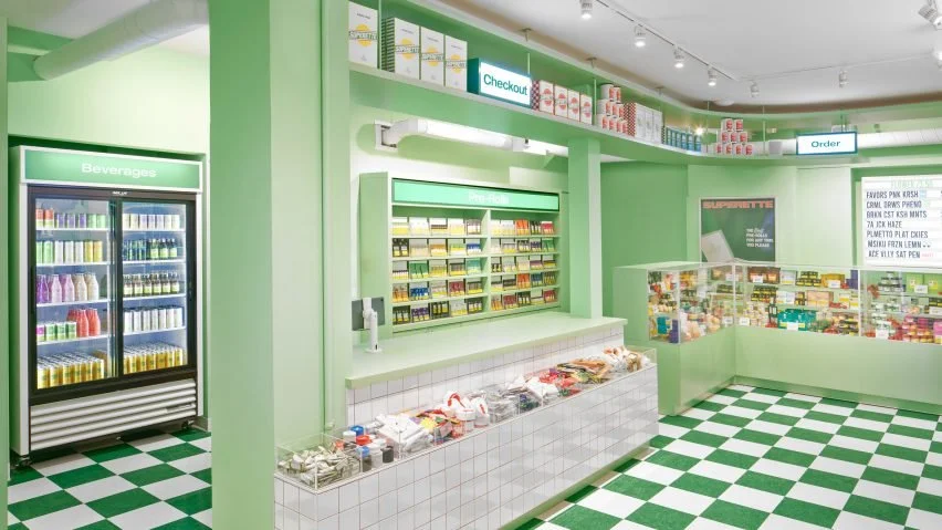 Inside a small convenience store with green walls and checkered green and white flooring, featuring beverage fridge, checkout counter with pre-rolls, and shelves with snacks and candies.