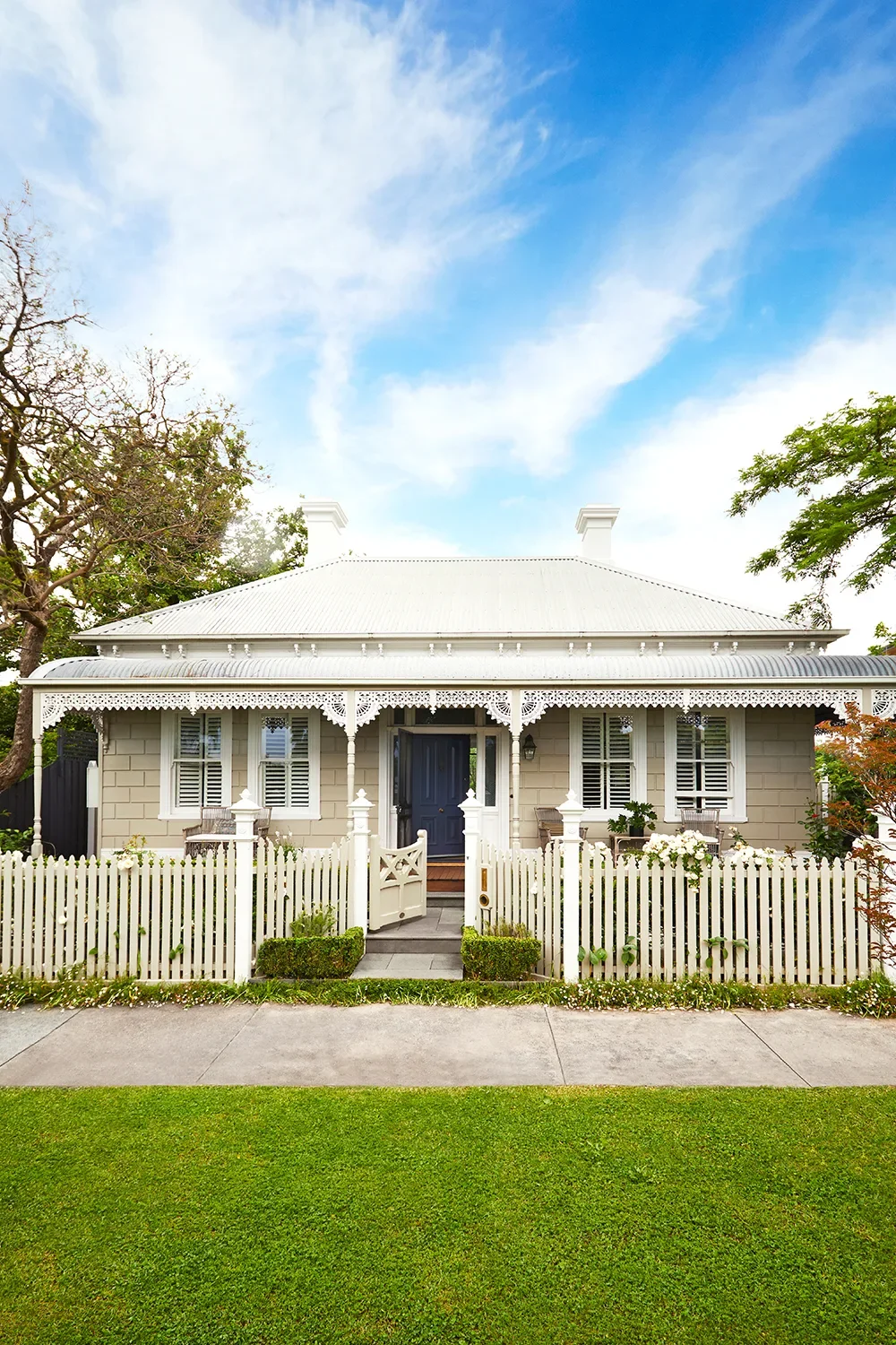 A charming, traditional house with a white picket fence, grey exterior walls, and white decorative trim. The front entrance has a dark door, and the house is surrounded by greenery and trees under a partly cloudy sky.