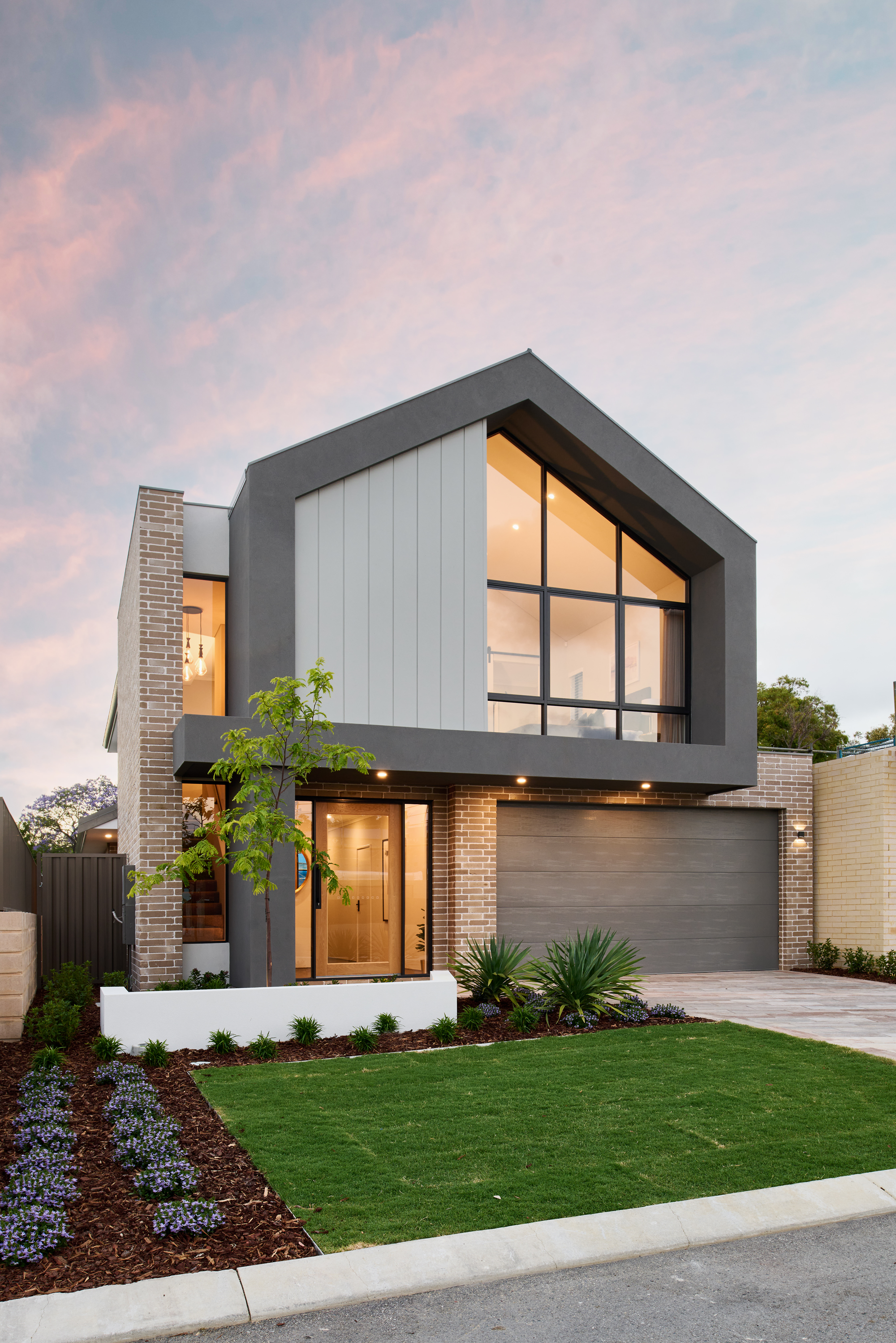 Modern residential house with large glass windows on the upper floor, brick and gray exterior, landscaped front yard with small trees, plants, and a green lawn, at sunset.