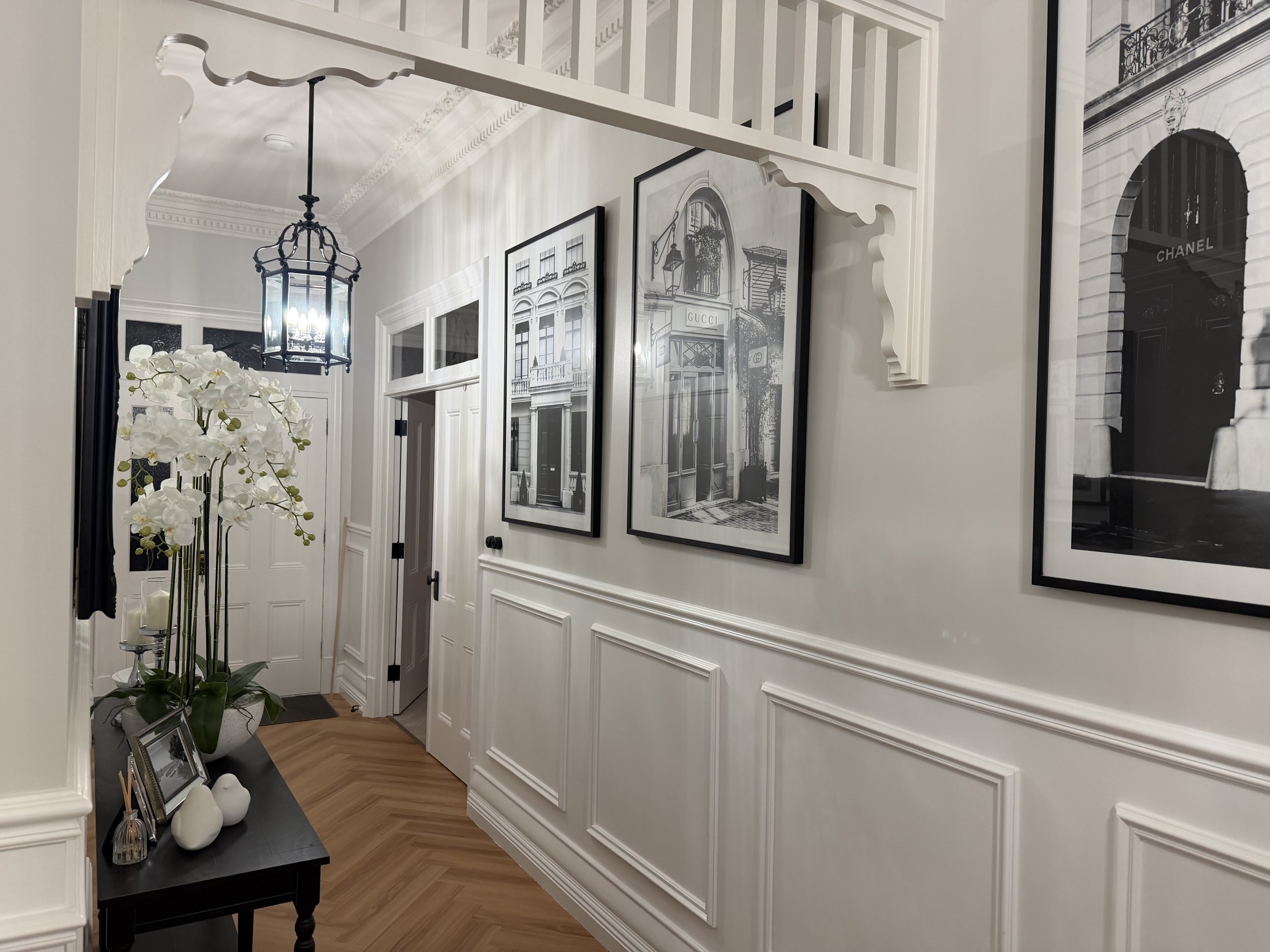 A hallway with white walls and framed black-and-white cityscape photographs. There is a black table with decorative white vases and picture frames, a large potted orchid, and a black chandelier hanging from the ceiling.