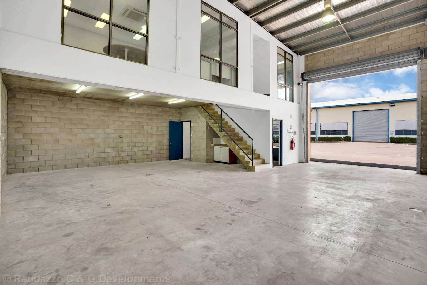 Empty industrial warehouse with large roll-up door open, concrete floor, brick and drywall walls, staircase leading to a mezzanine office area with windows