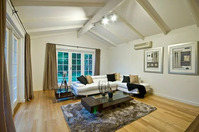 Living room with white sectional sofa, glass coffee table, wooden floors, large windows with curtains, and framed artwork on the wall.
