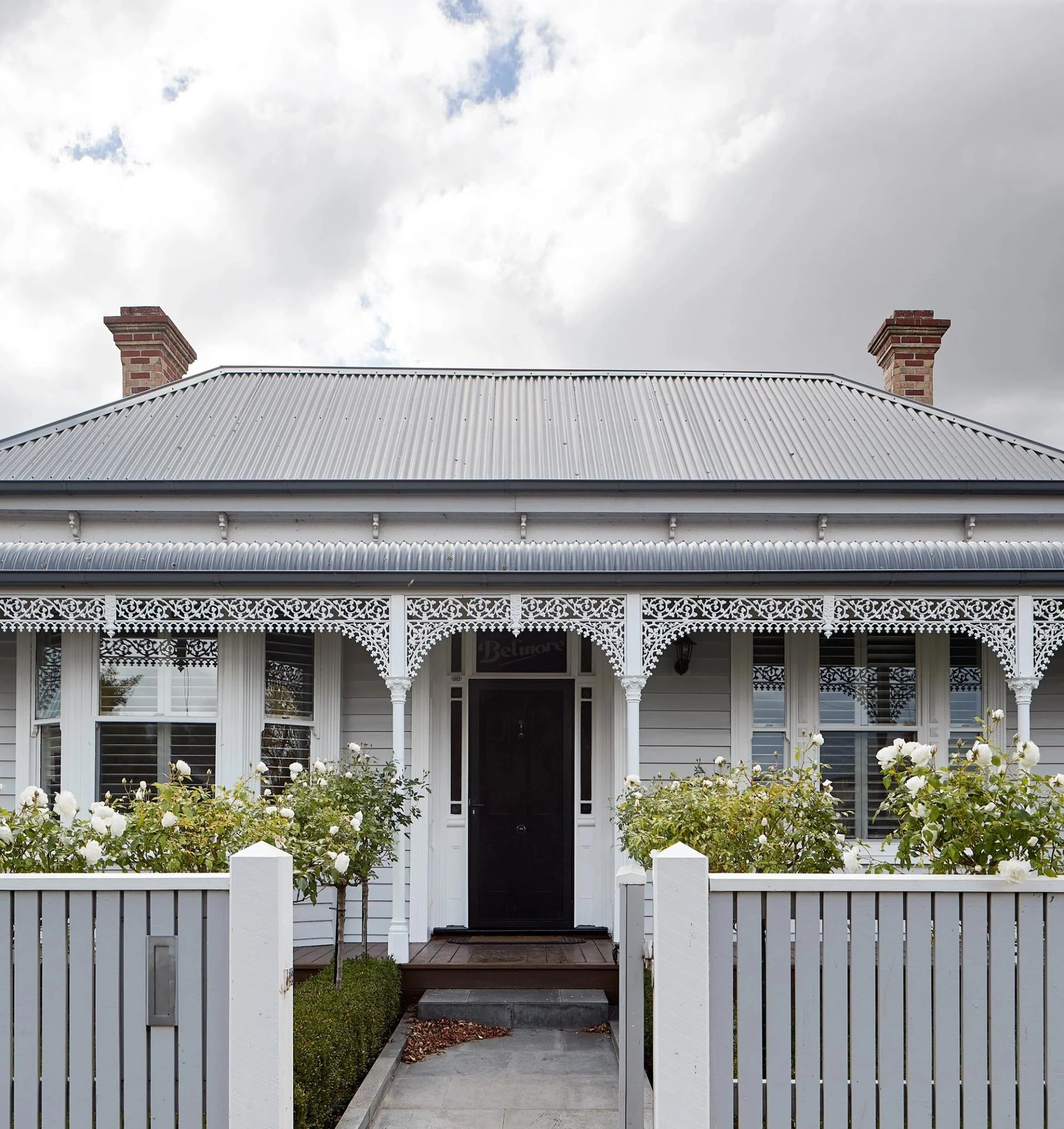 Front view of a white house with a porch, two brick chimneys, a metal roof, and a white picket fence with two small trees in front, under a cloudy sky.