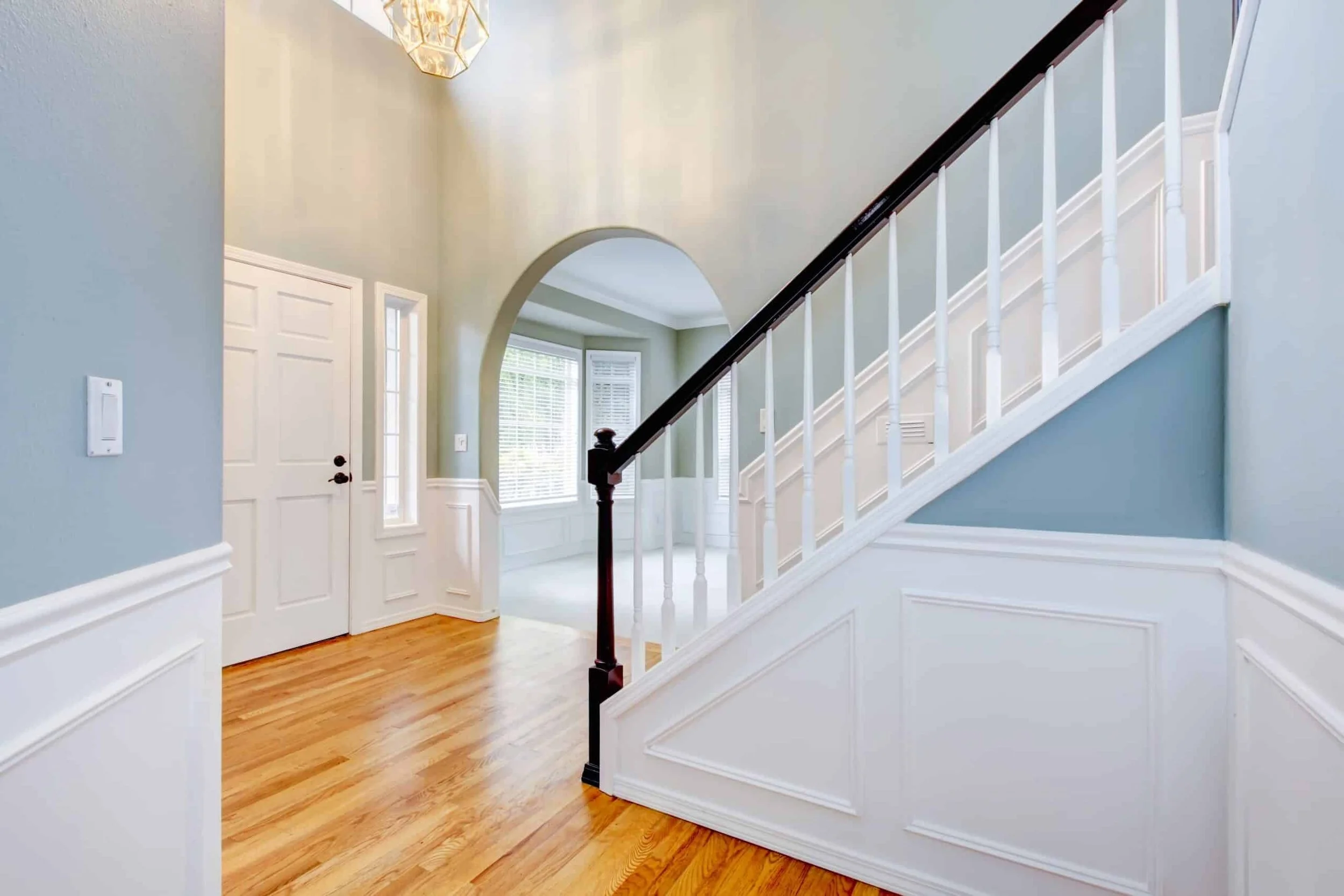 Interior view of a house entryway with hardwood floors, a staircase with white balusters and a dark wooden handrail, a white door, large windows, and an arched doorway with a ceiling light fixture.
