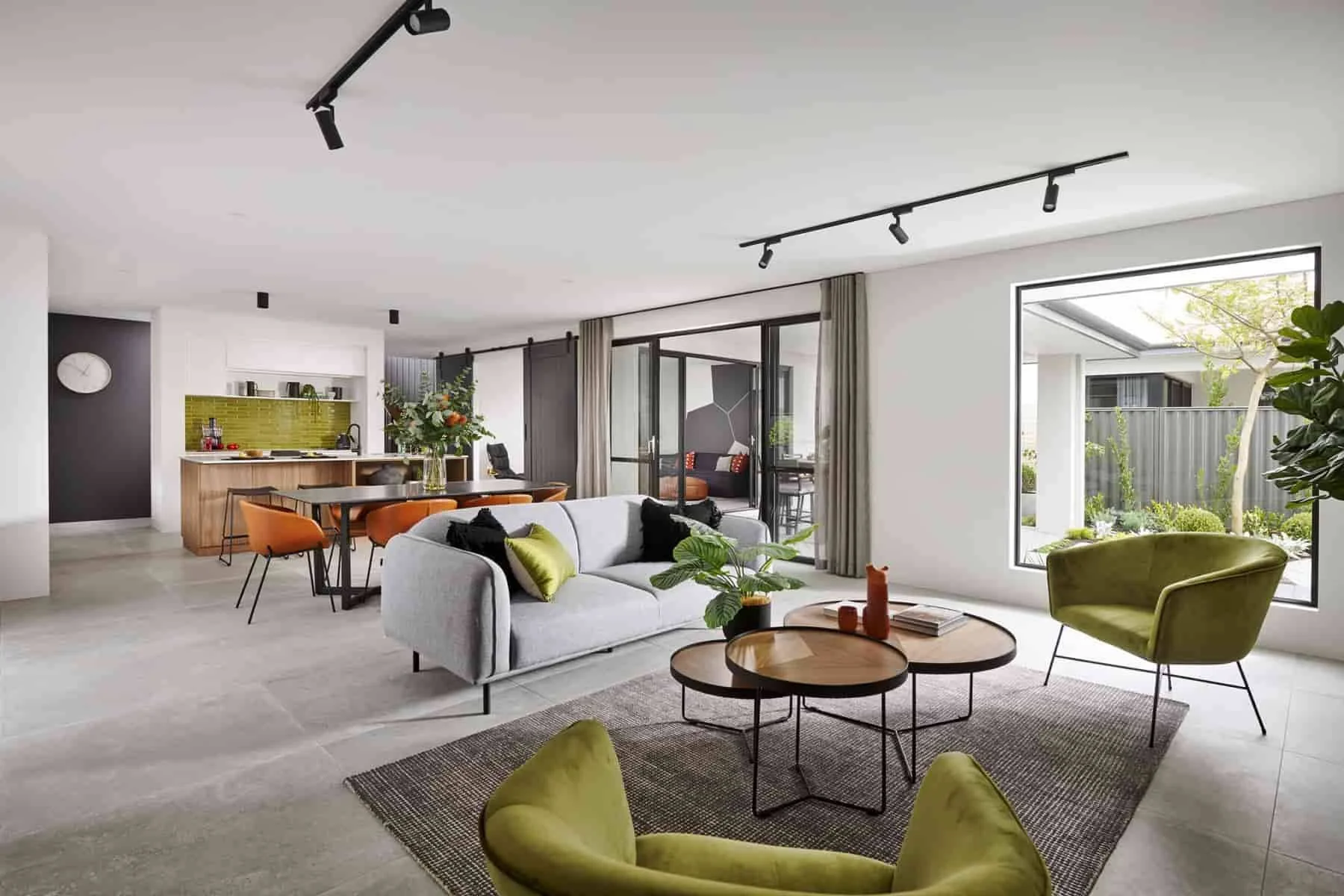 Modern open-plan living room with white walls, gray tiled floor, and large windows. Contains a white sofa with black and green cushions, a round wooden coffee table, and green armchairs. The space is decorated with houseplants and contemporary decor. In the background, a kitchen with a yellow backsplash and dining table with orange chairs is visible.