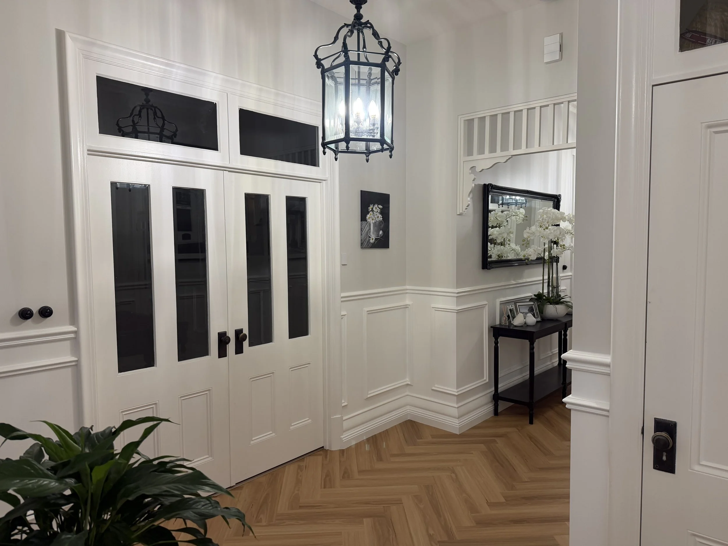 An elegant entryway with white double doors featuring glass panels, a black chandelier hanging above, a small black table with decorative items including a plant and picture frames, and a wall mirror reflecting flowers.